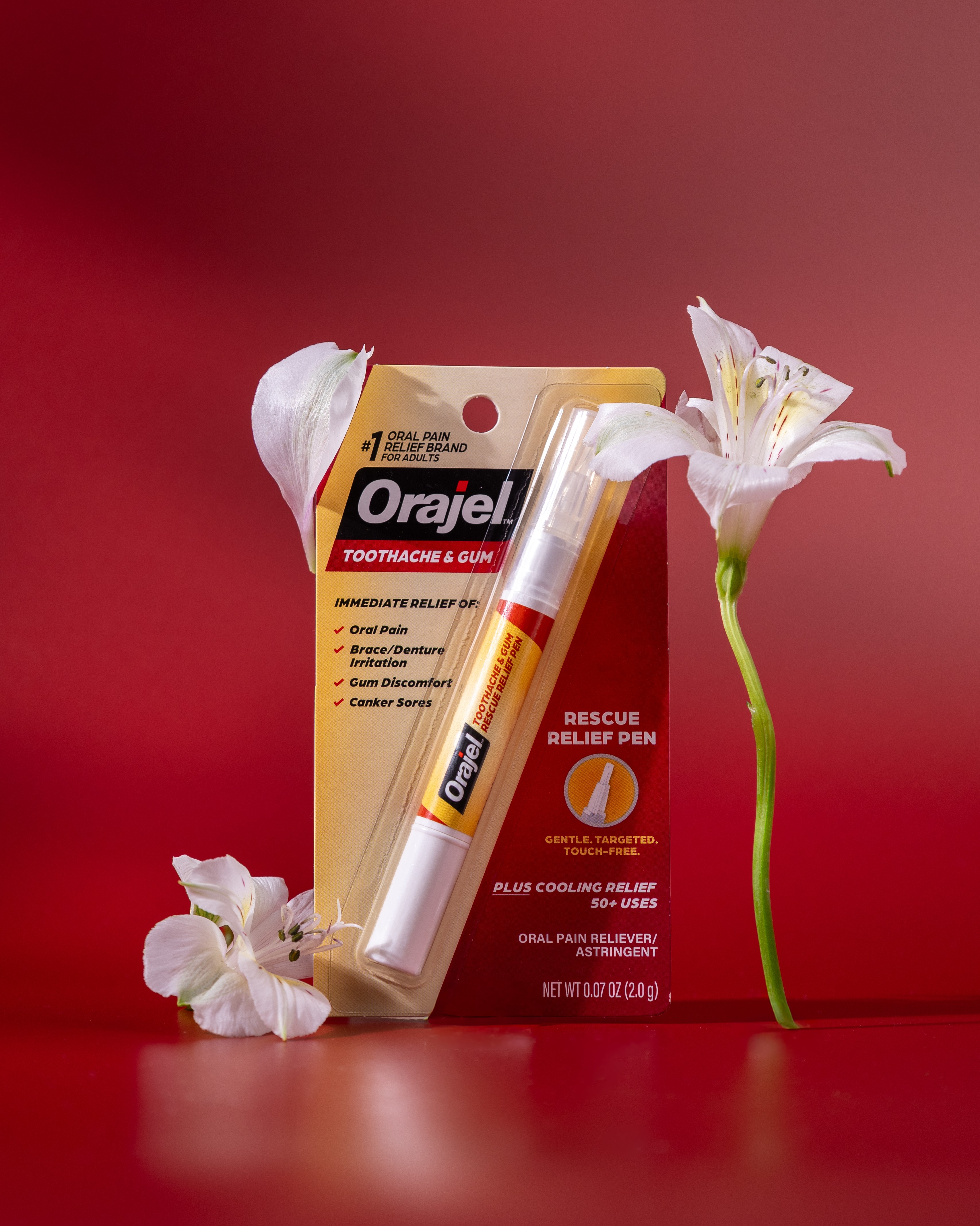 A white and yellow Orajel toothache and gum relief pen in packaging, with white flowers on a red background.