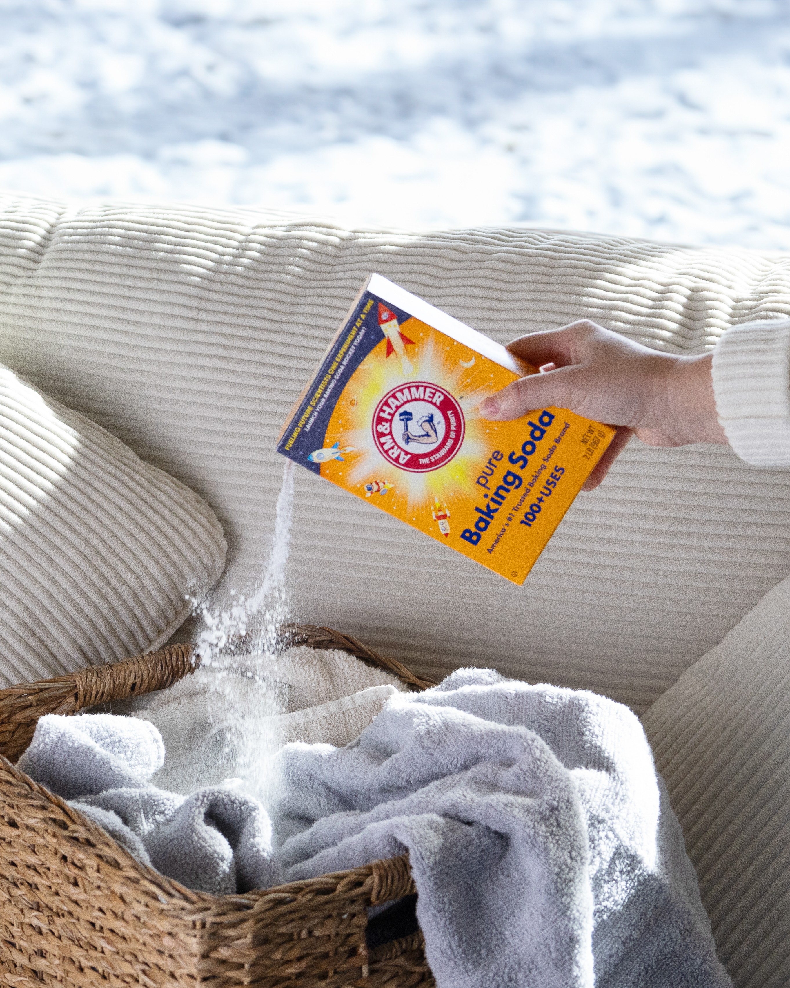 Person pouring baking soda from a box onto a blanket on a sofa, with pillows and a window in the background.