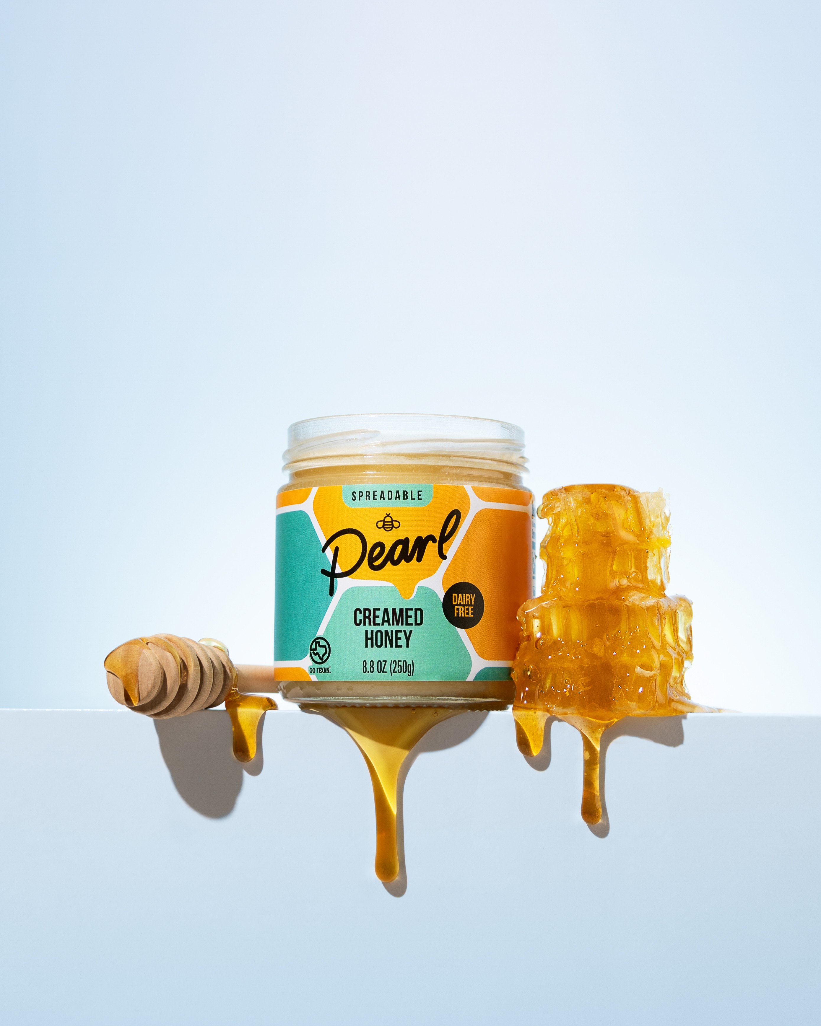Open jar of creamed honey with honeycomb and honey dipper on a light blue background.