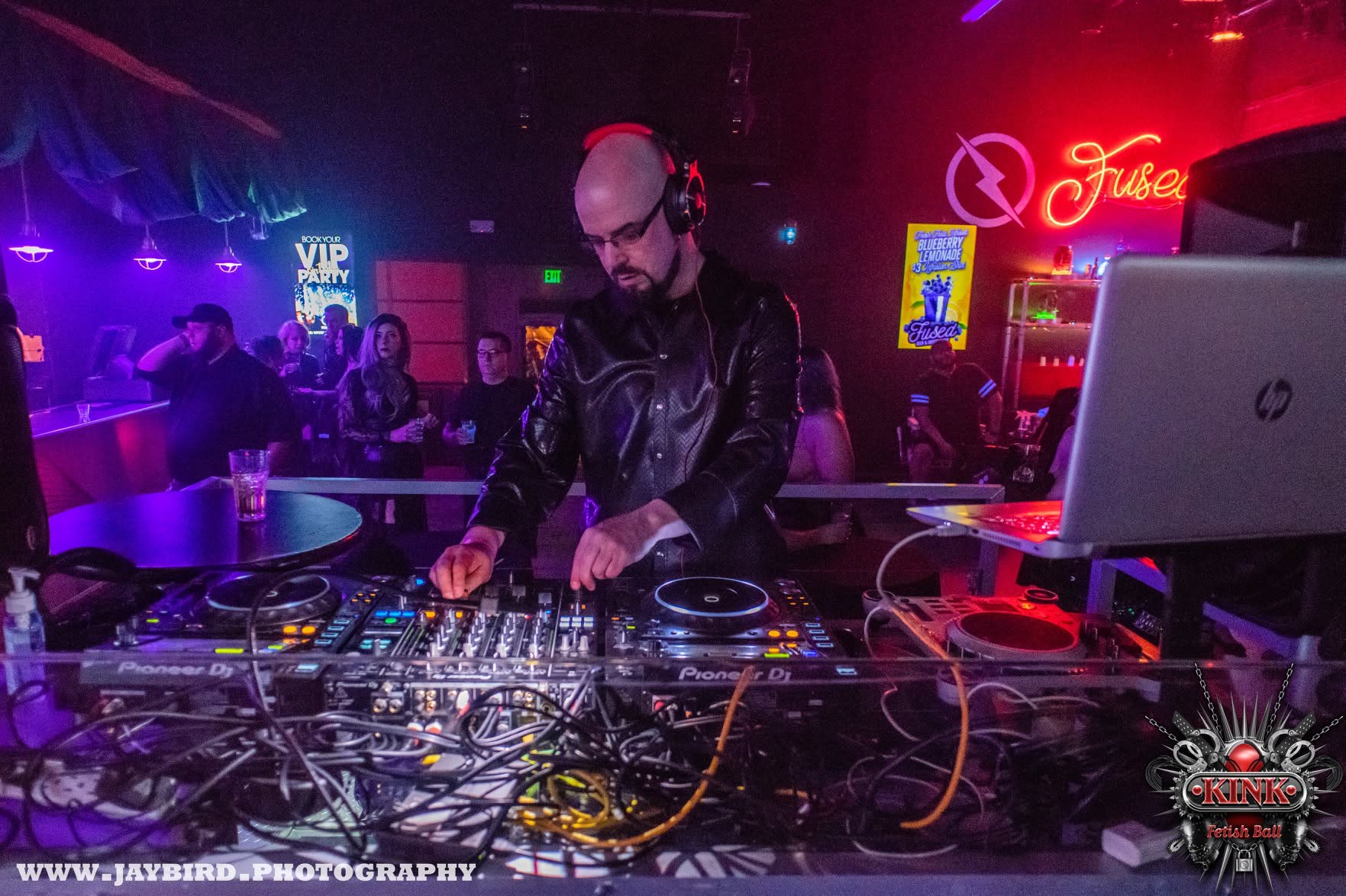 DJ with headphones operating turntables in a nightclub with colorful lights and a crowd in the background.