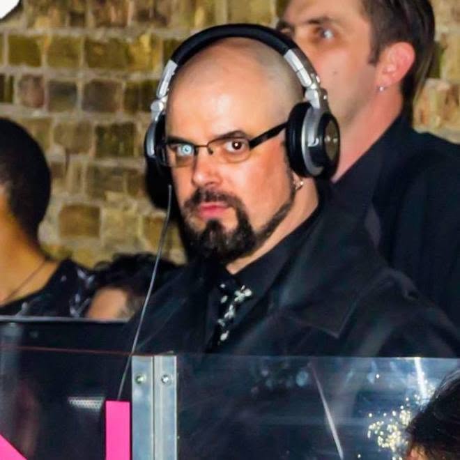 Man with glasses and headphones at a social event, with a brick wall background.