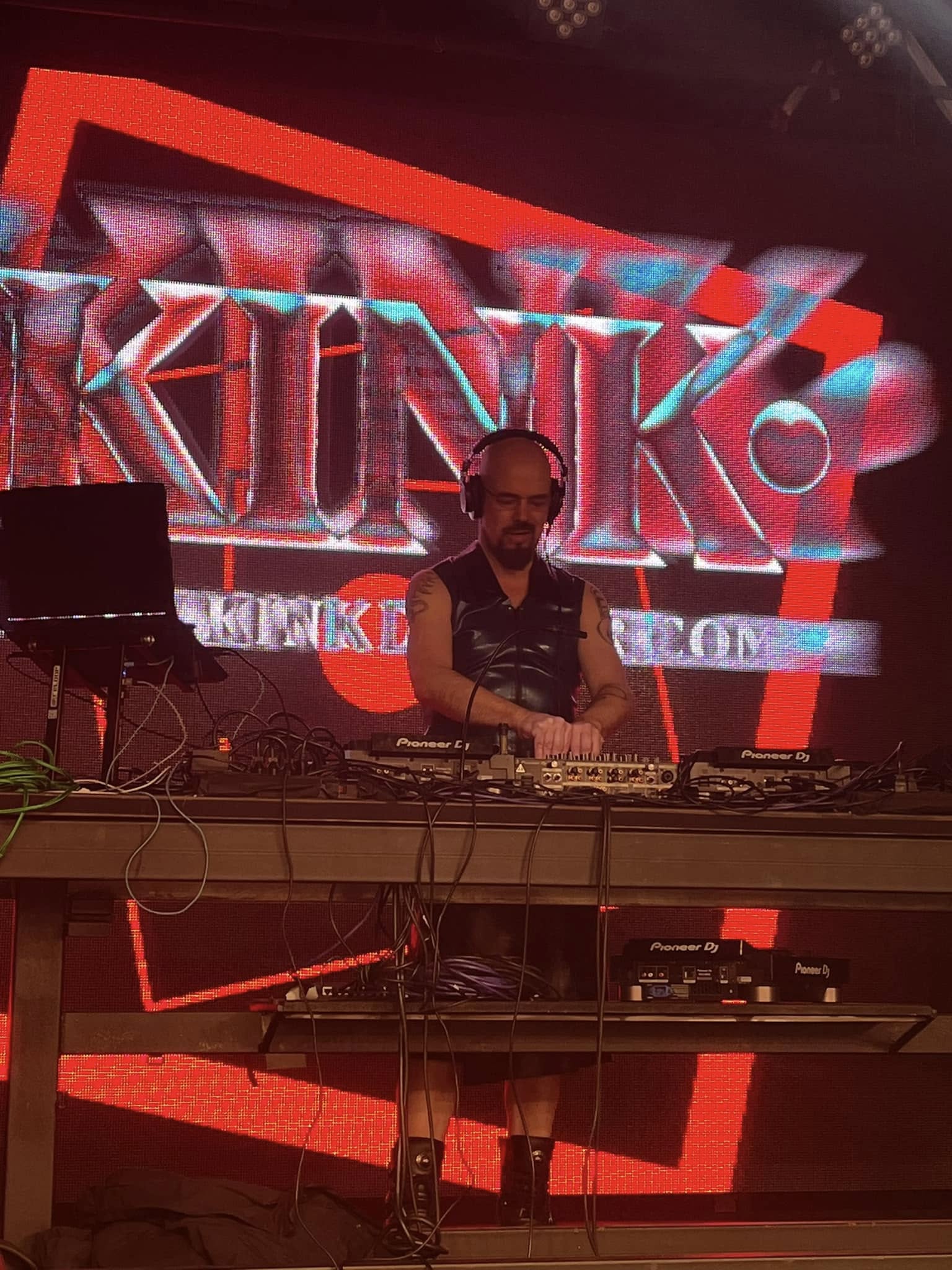 DJ performing on stage with a large colorful digital display behind him, electronic equipment on table, dark background.