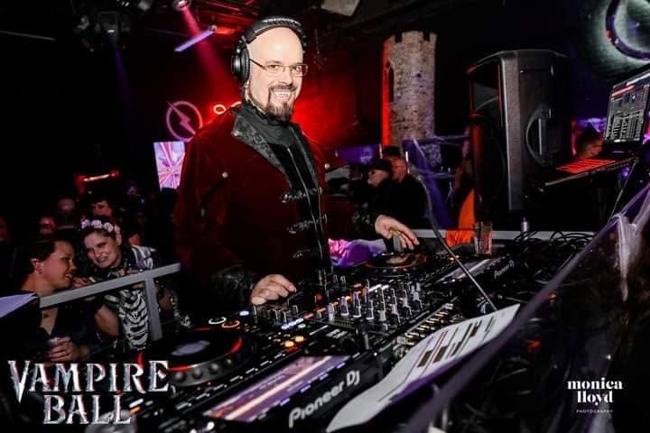 DJ with headphones at a nightclub, crowd in background, red and purple lighting, equipment on table, smiling man, dark clothing
