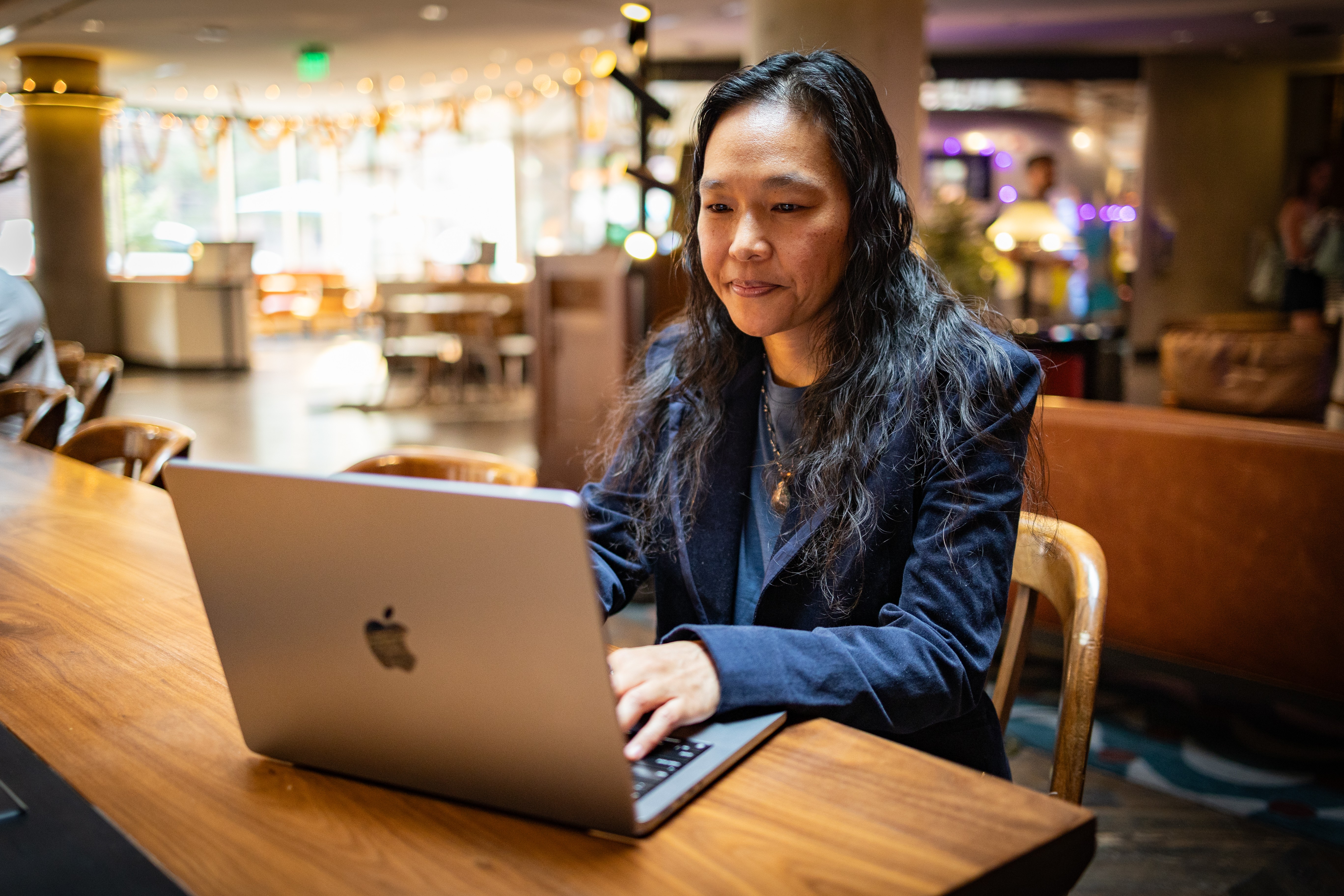 Woman with long dark hair working on a laptop at a wooden table in a cozy cafe or restaurant.