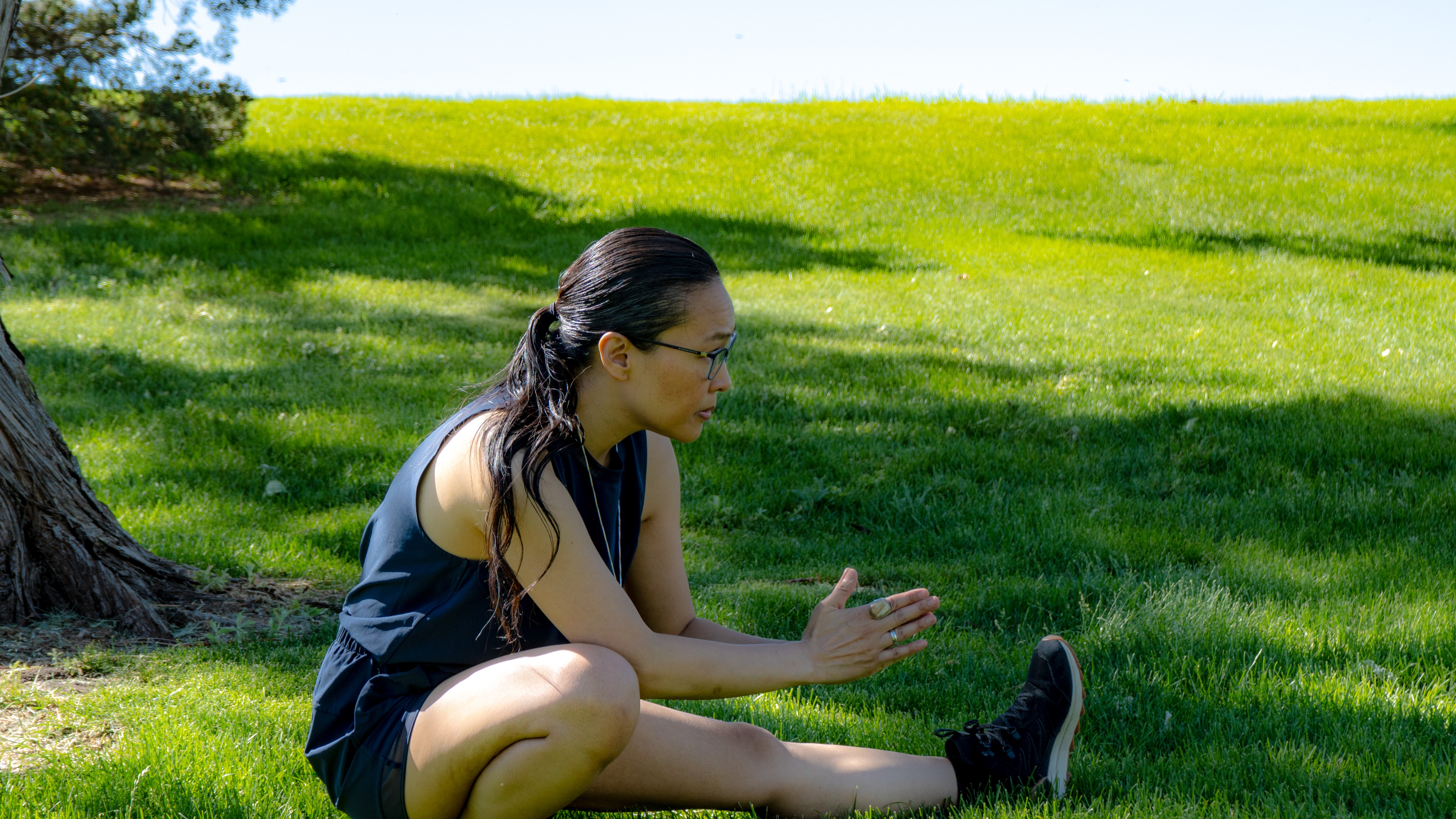 Woman sitting on grass with legs extended, leaning forward, wearing glasses and a dark sleeveless top, outdoors on a sunny day.