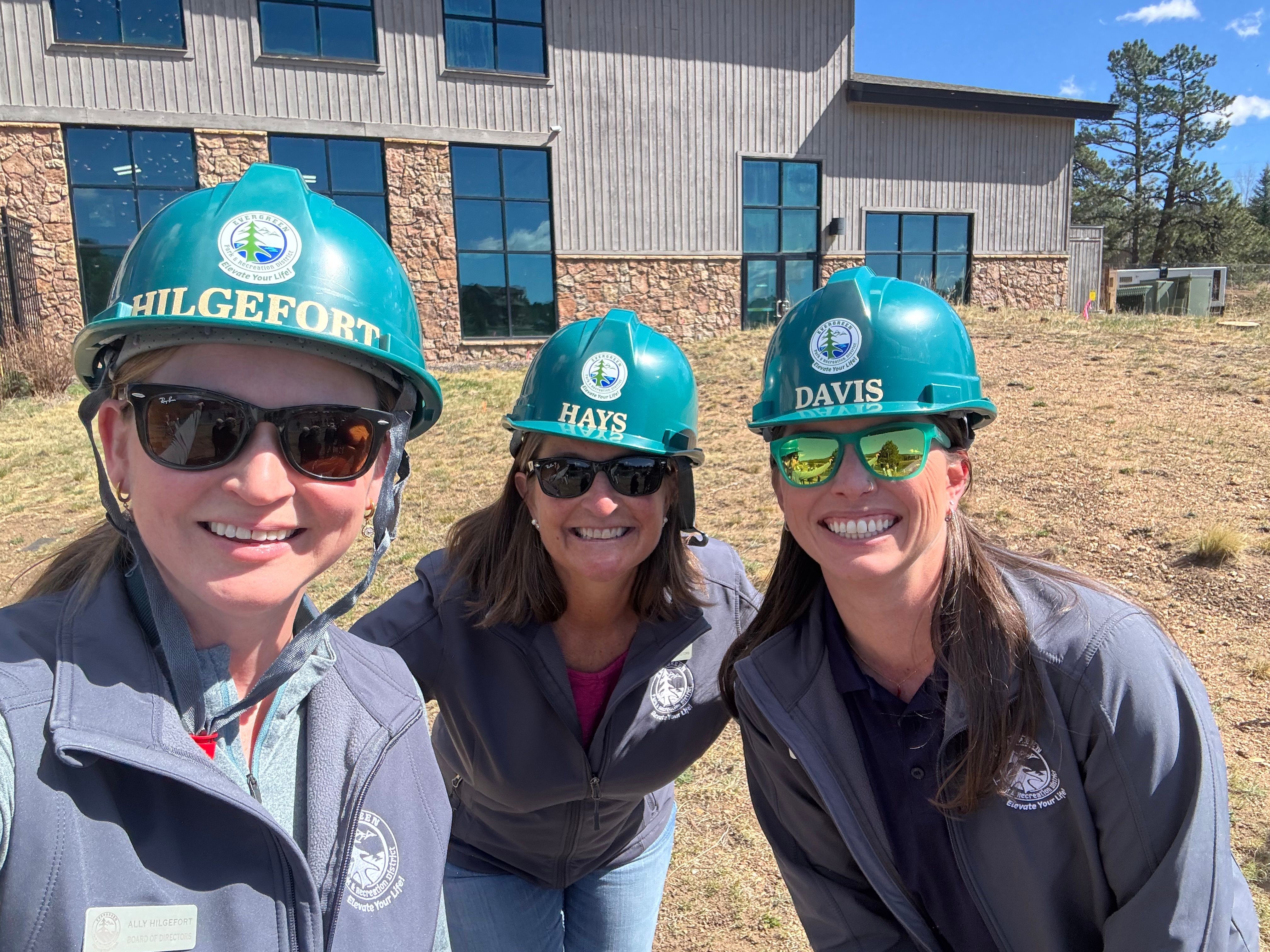 Three people wearing blue helmets and sunglasses stand outdoors in front of a building, smiling at the camera.