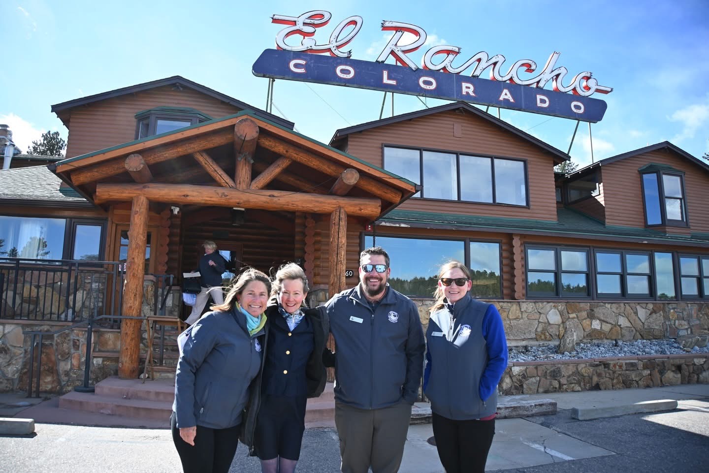 Four people stand in front of a wooden building with a sign that reads 'El Rancho Colorado' against a blue sky.