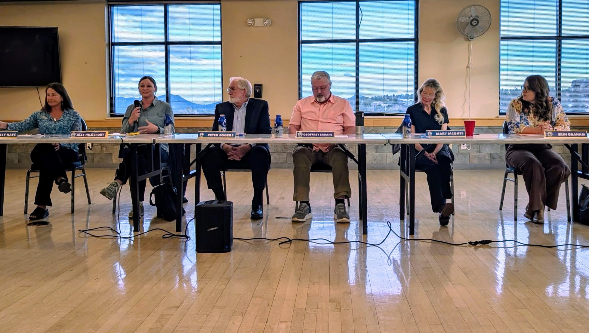 Six people seated at a long table in a room with large windows and a wooden floor.