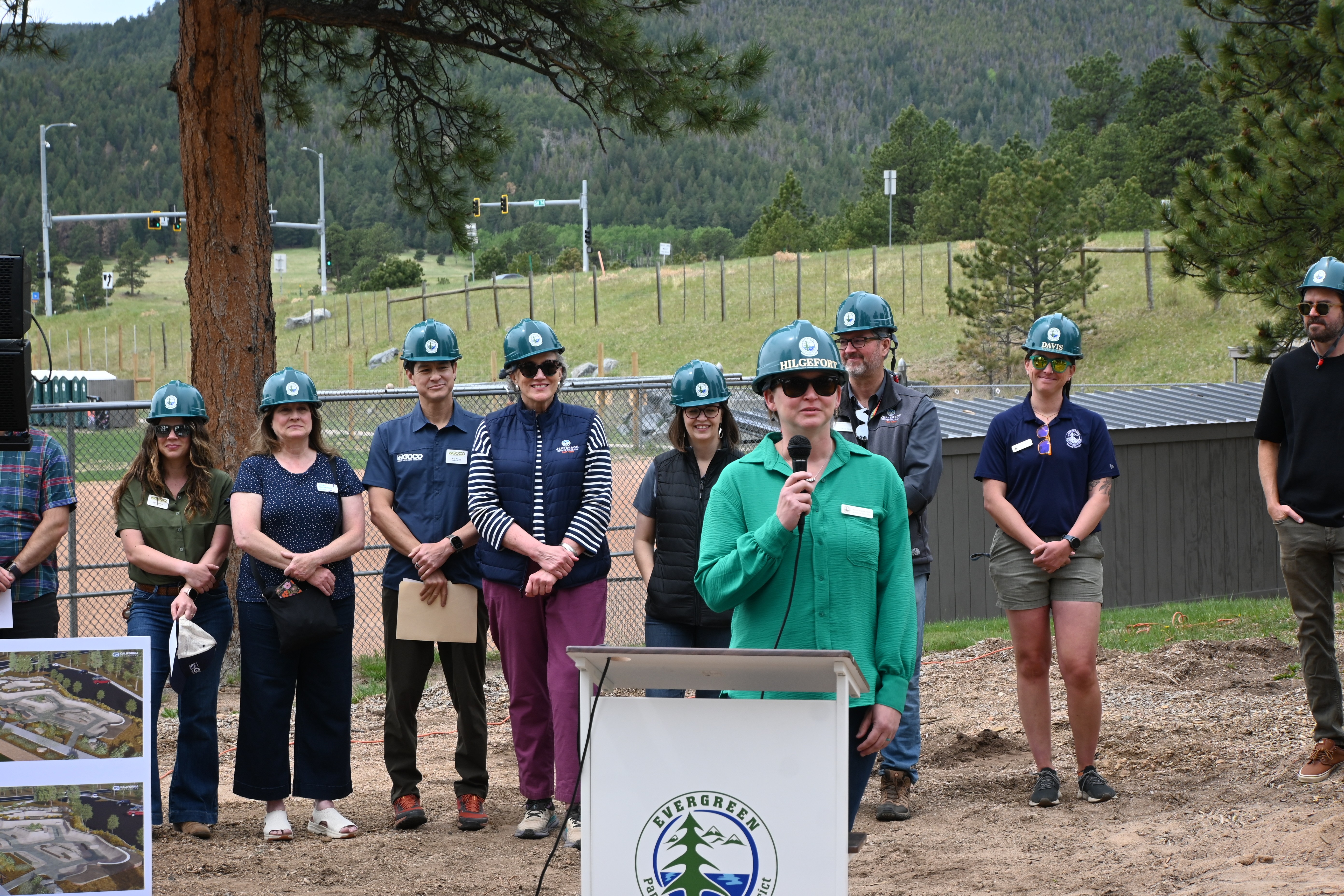 Group of people wearing helmets standing outdoors, with a woman speaking into a microphone in front of a sign, trees and a fence in background.