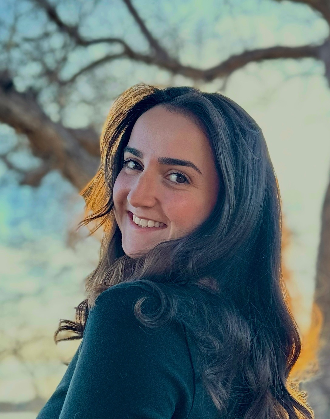 Young woman with long dark hair smiling outdoors, background of trees and sky, sunlight illuminating her face.
