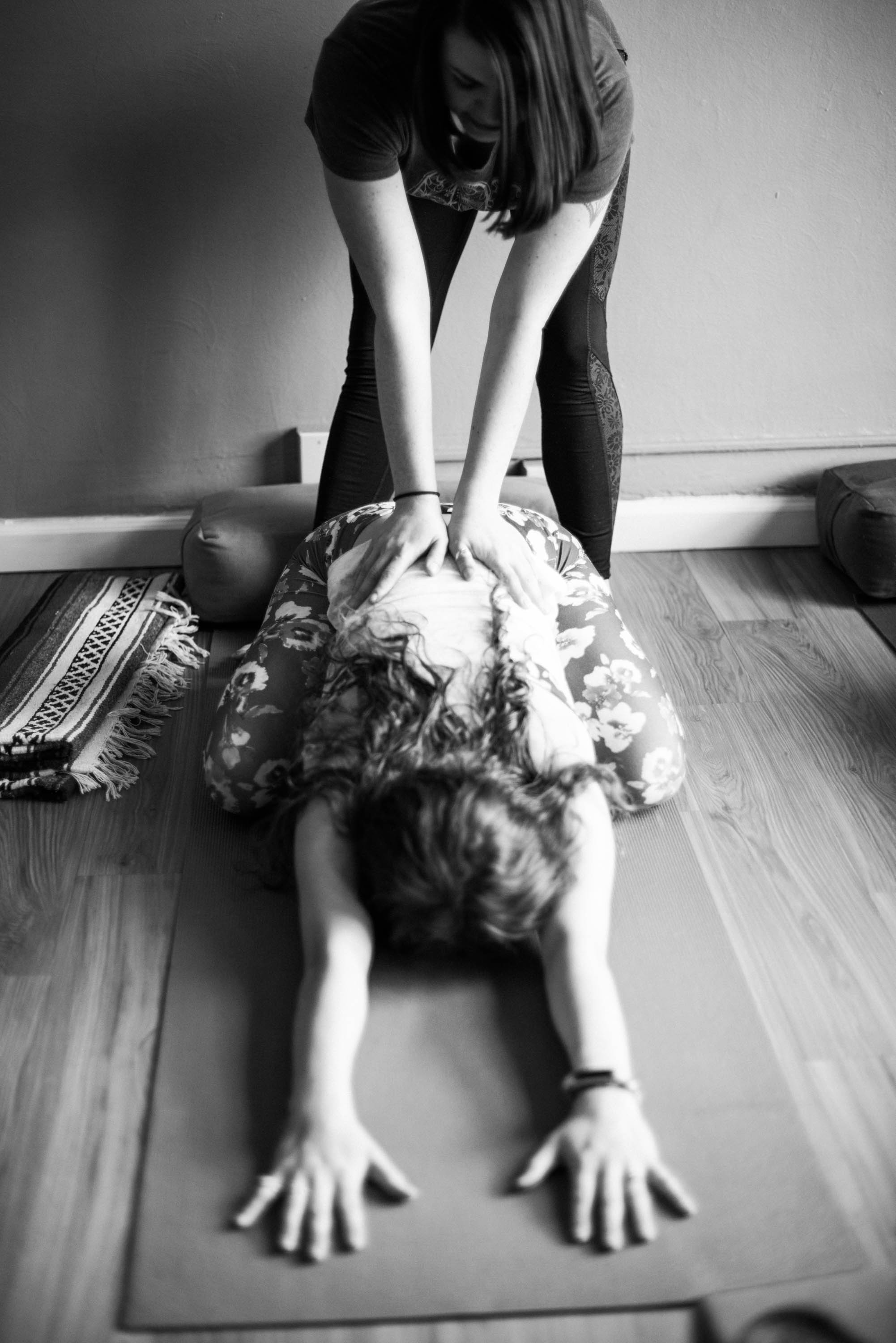 Person performing yoga pose on mat with child lying face down, hands extended forward, in a room with wooden floor.