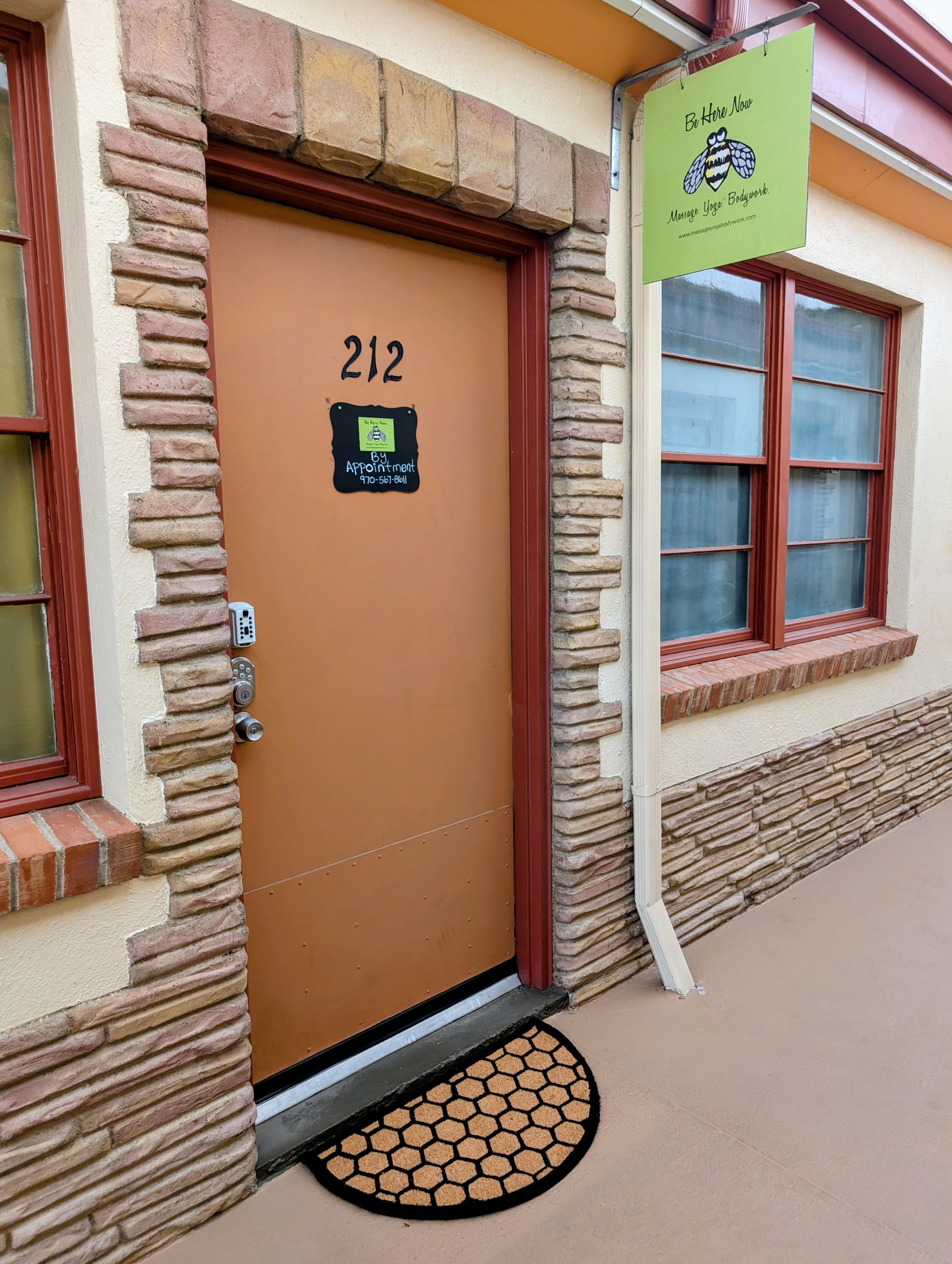 Brown door with the number 212, surrounded by brick and stone, with a black doormat in front, and a green sign above.