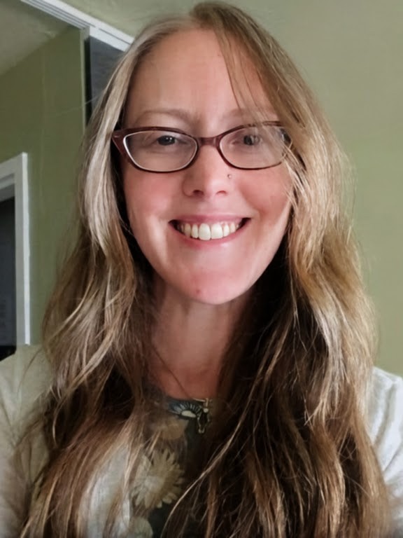 Woman with long wavy hair, glasses, smiling, indoors with green wall background.