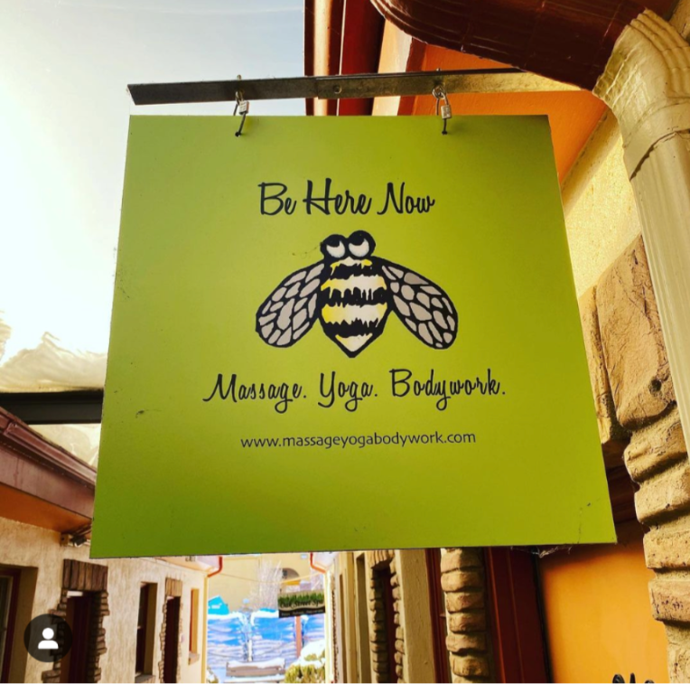 Green sign with bee illustration and text about massage, yoga, and bodywork, hanging indoors near a window.