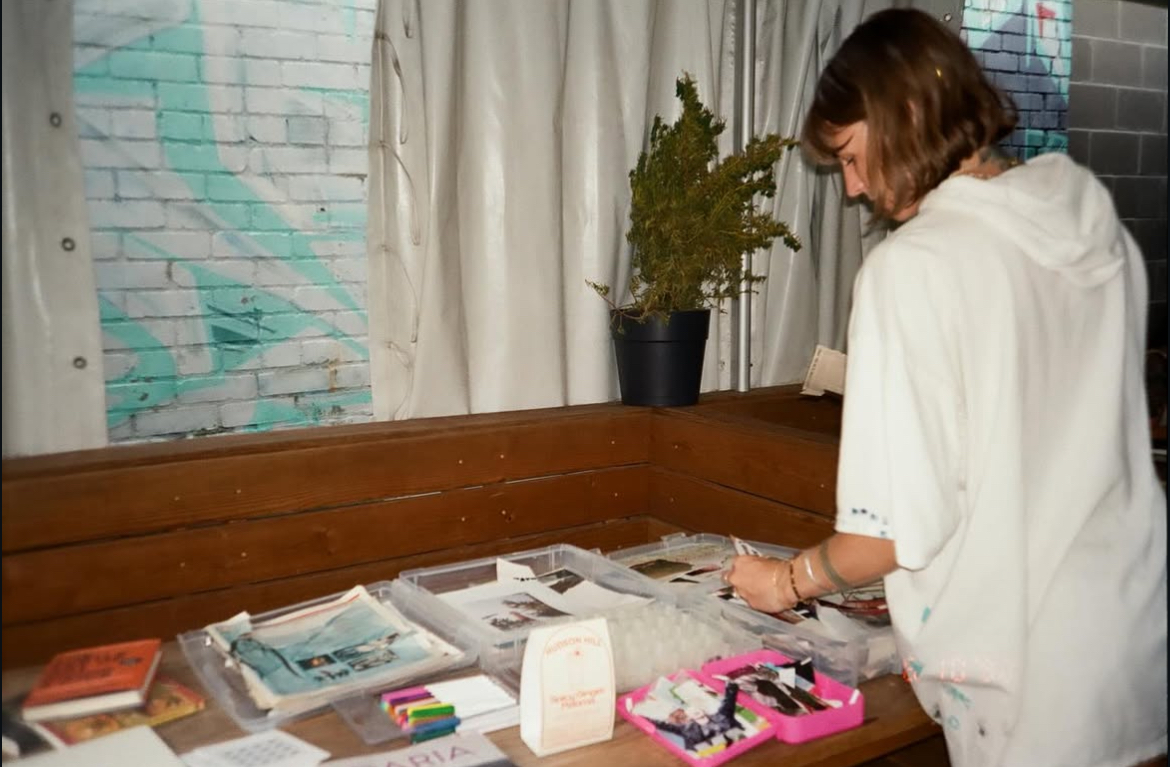 Person with shoulder-length hair looking at a table with papers and stationery, near a window with curtains.
