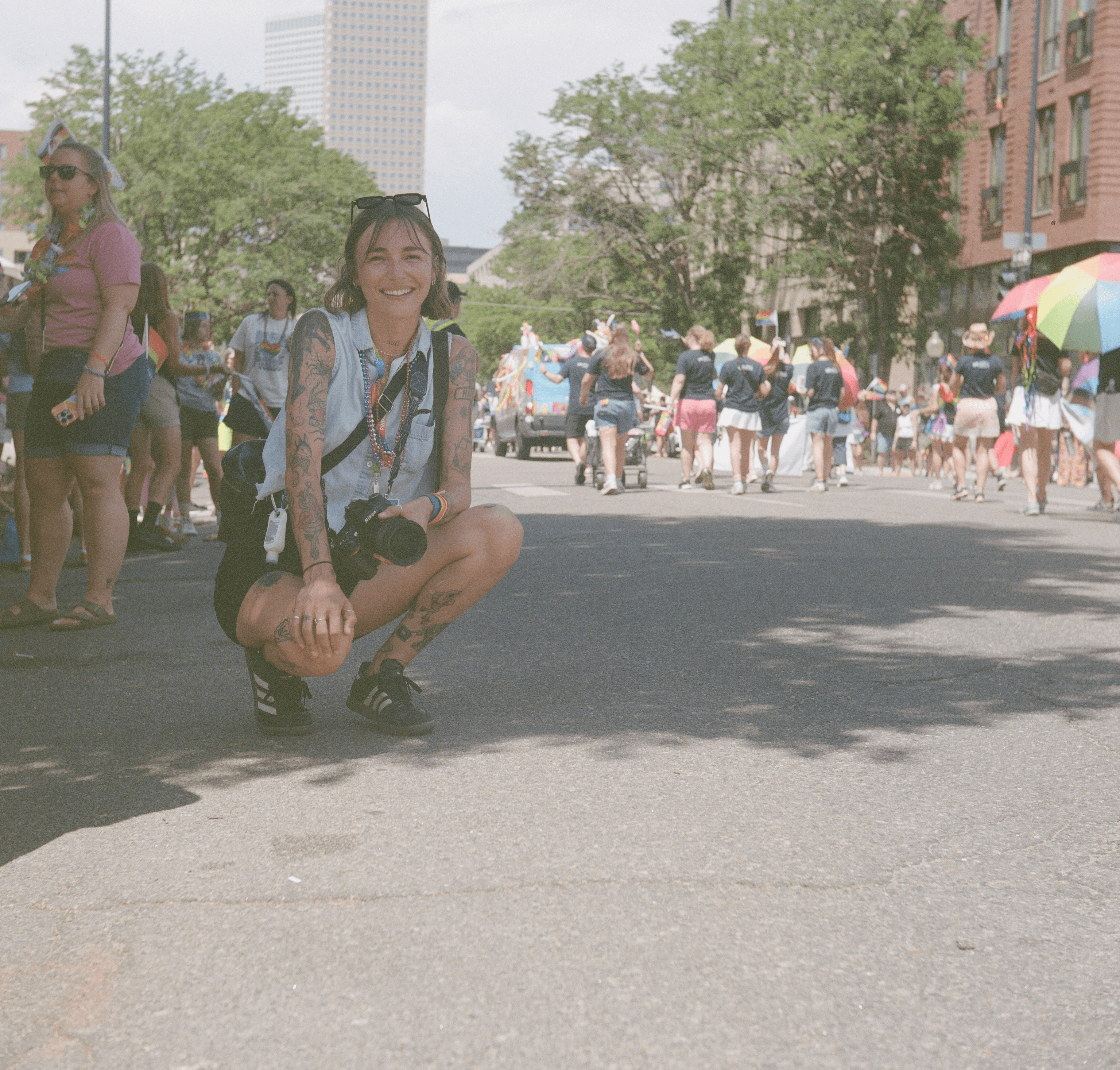 Young woman crouching on street with a crowd and trees in background, holding a camera, smiling, sunny day.