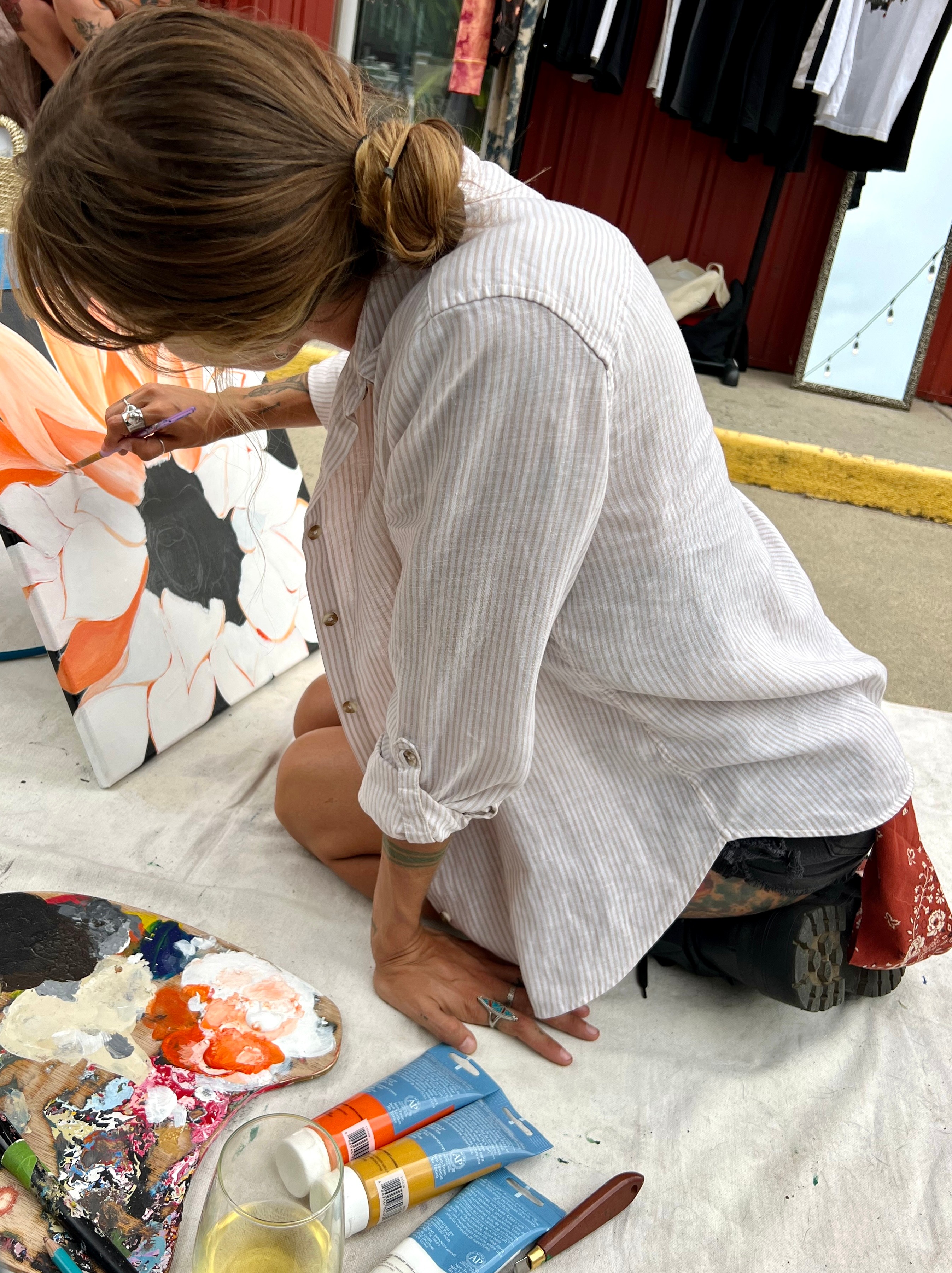 Person kneeling on the ground, painting on a canvas with a floral design, surrounded by paint supplies.