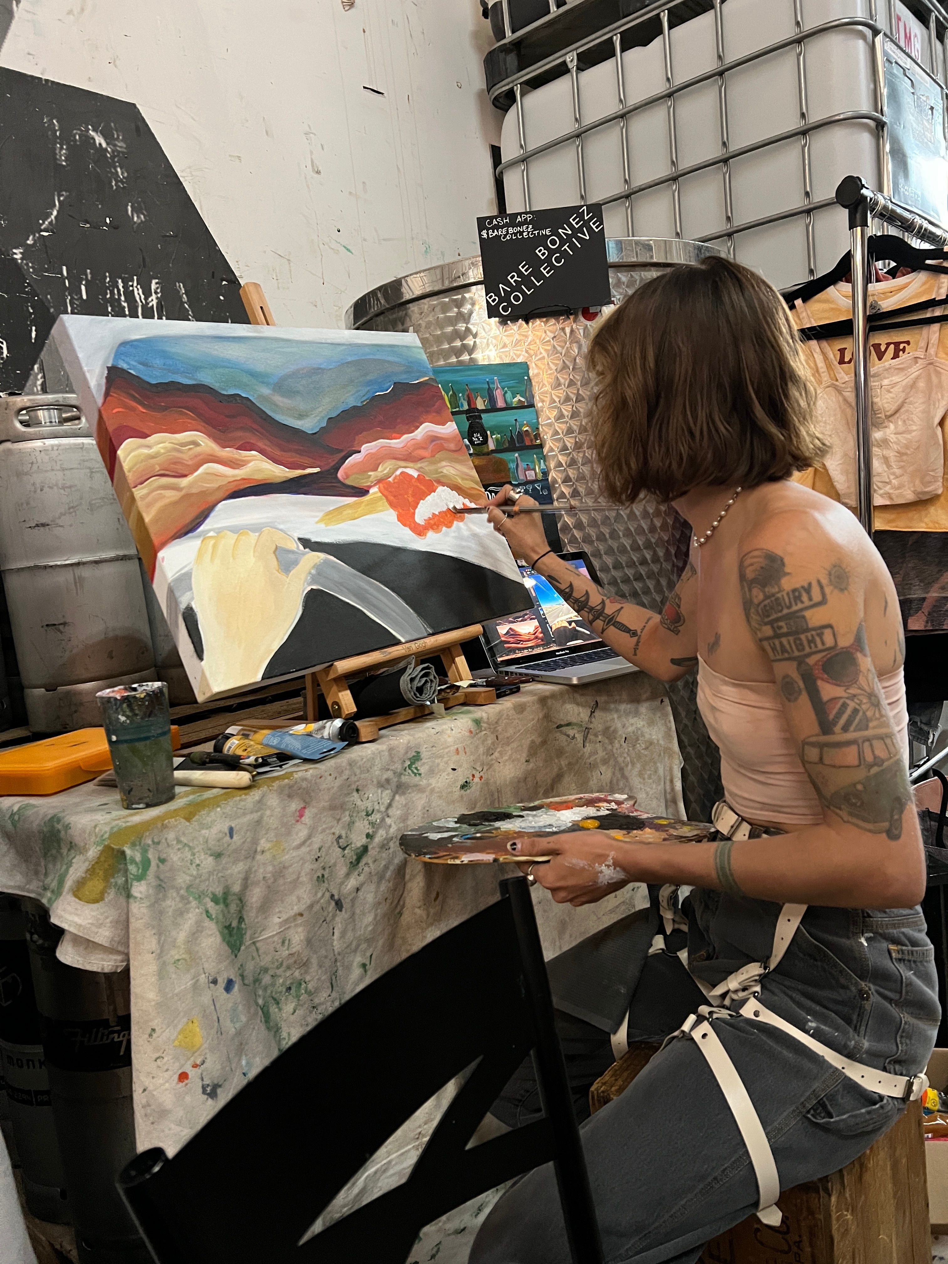 Person with tattoos painting a landscape on canvas in an art studio.