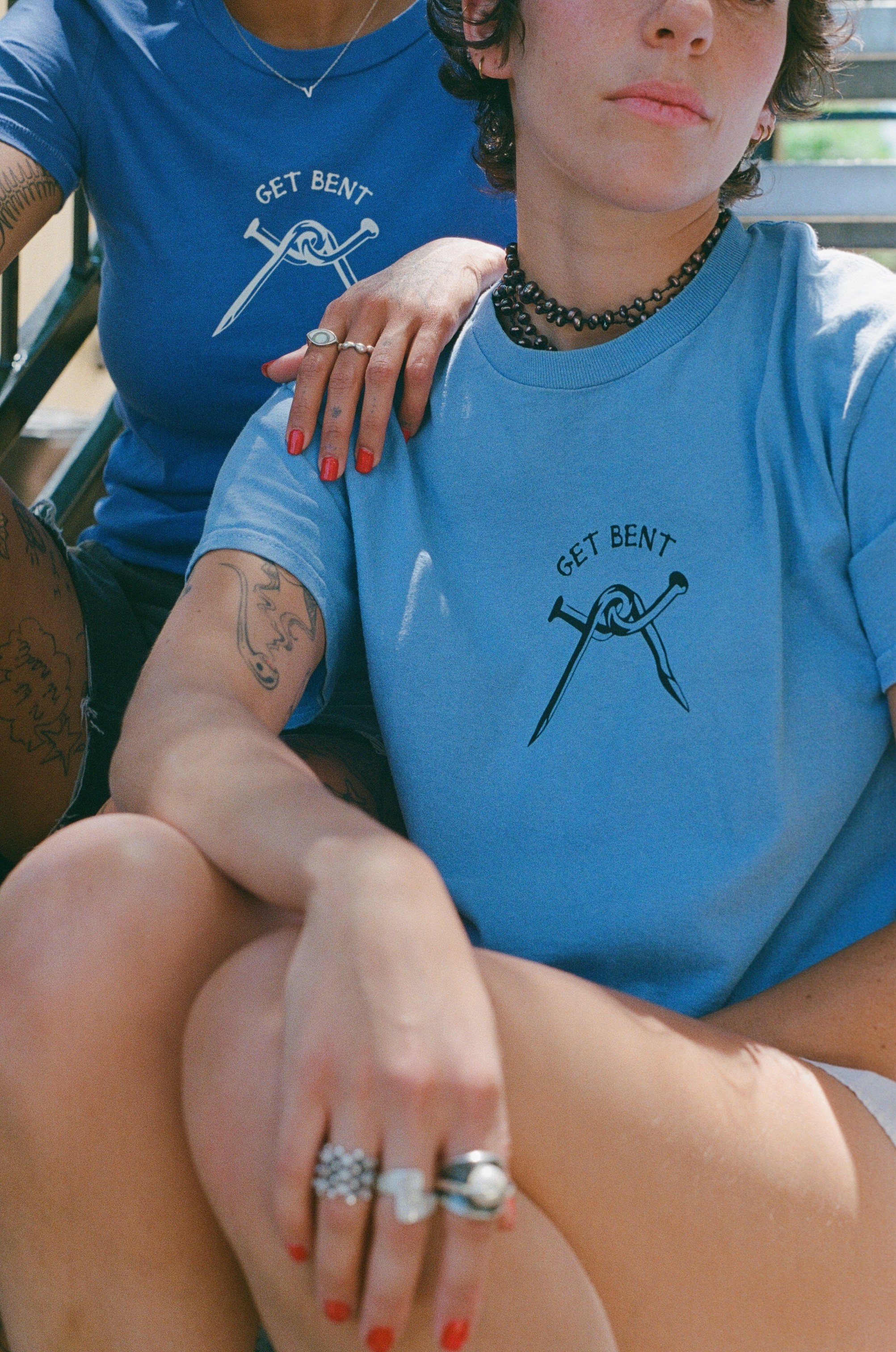 Two people wearing blue shirts with a scissors graphic and text, one with tattoos and rings, sitting close together.