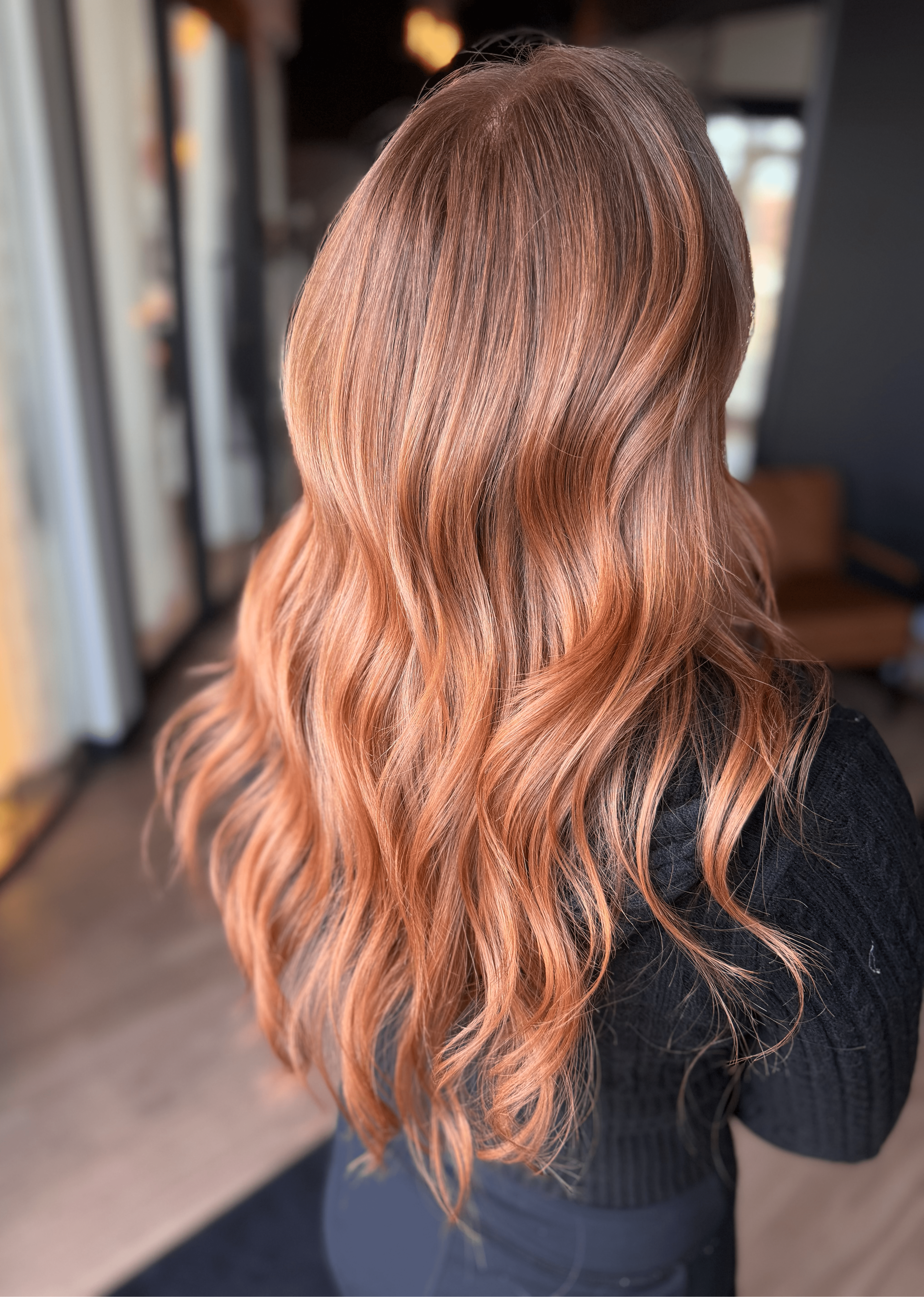 Back view of a woman with long, wavy, reddish-brown hair indoors, wearing a black top.