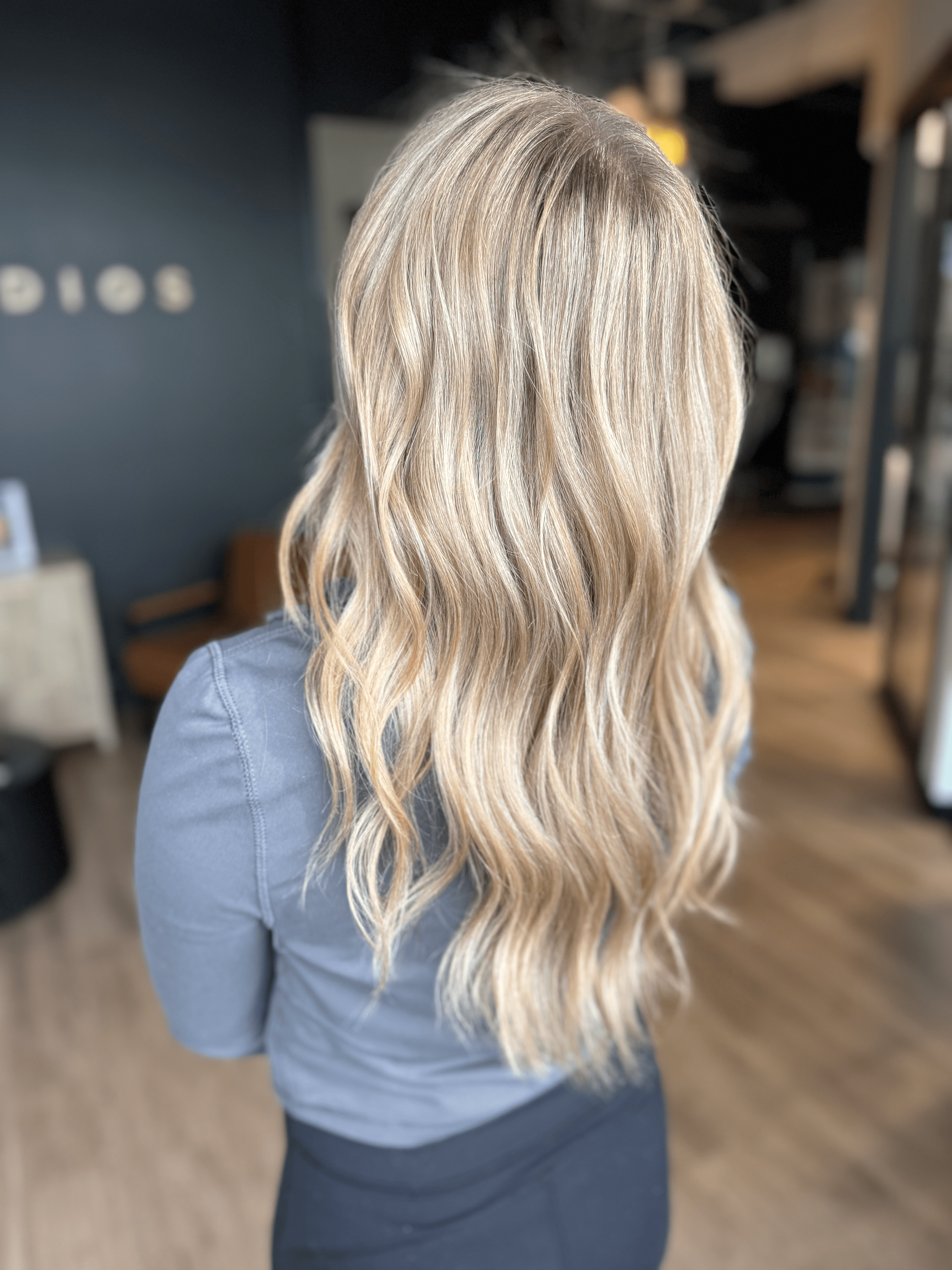 Back view of a woman with long, wavy blonde hair wearing a blue top in an indoor setting.