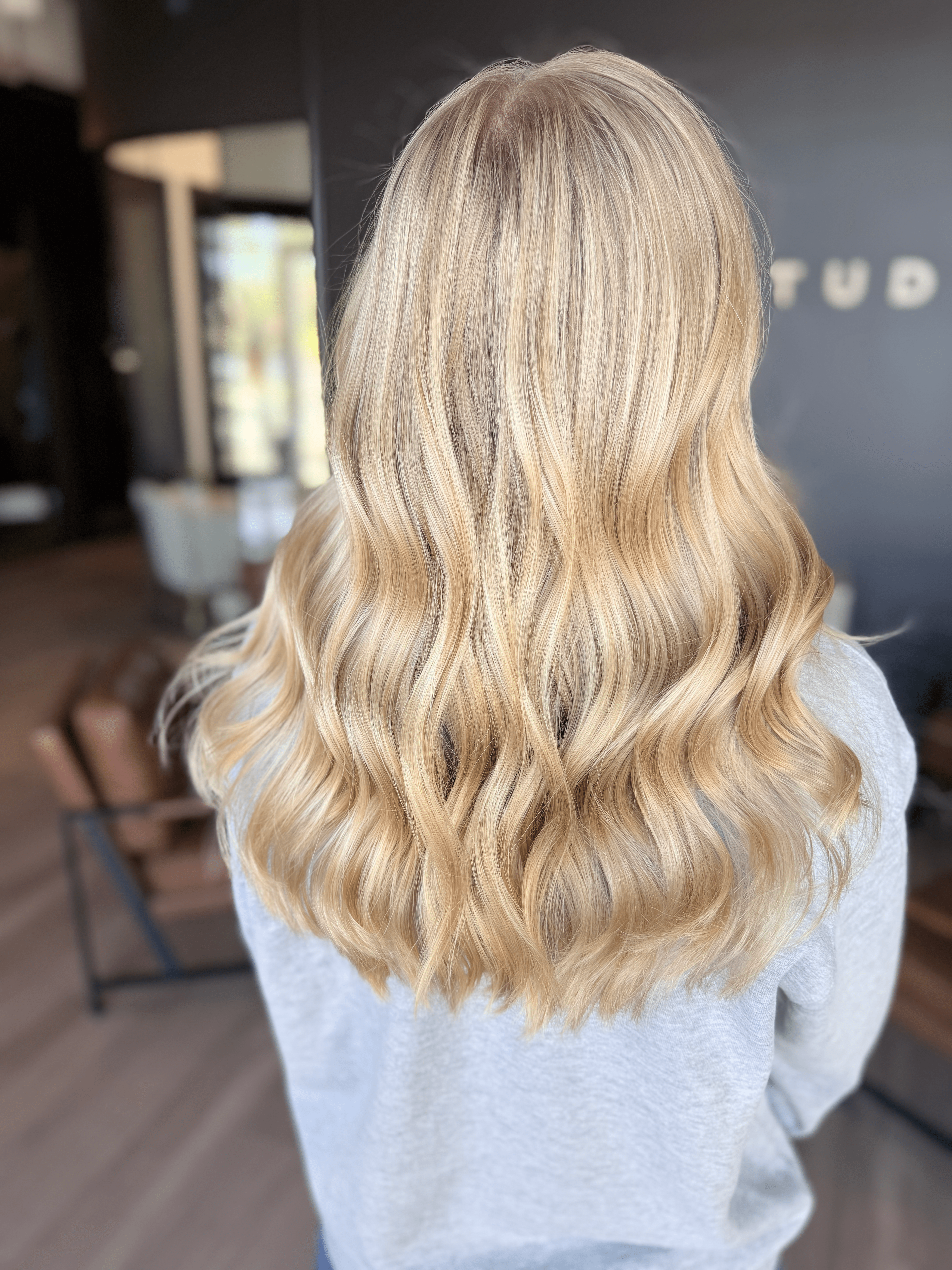Back view of a person with long, wavy blonde hair, wearing a light gray top, in a modern indoor setting.