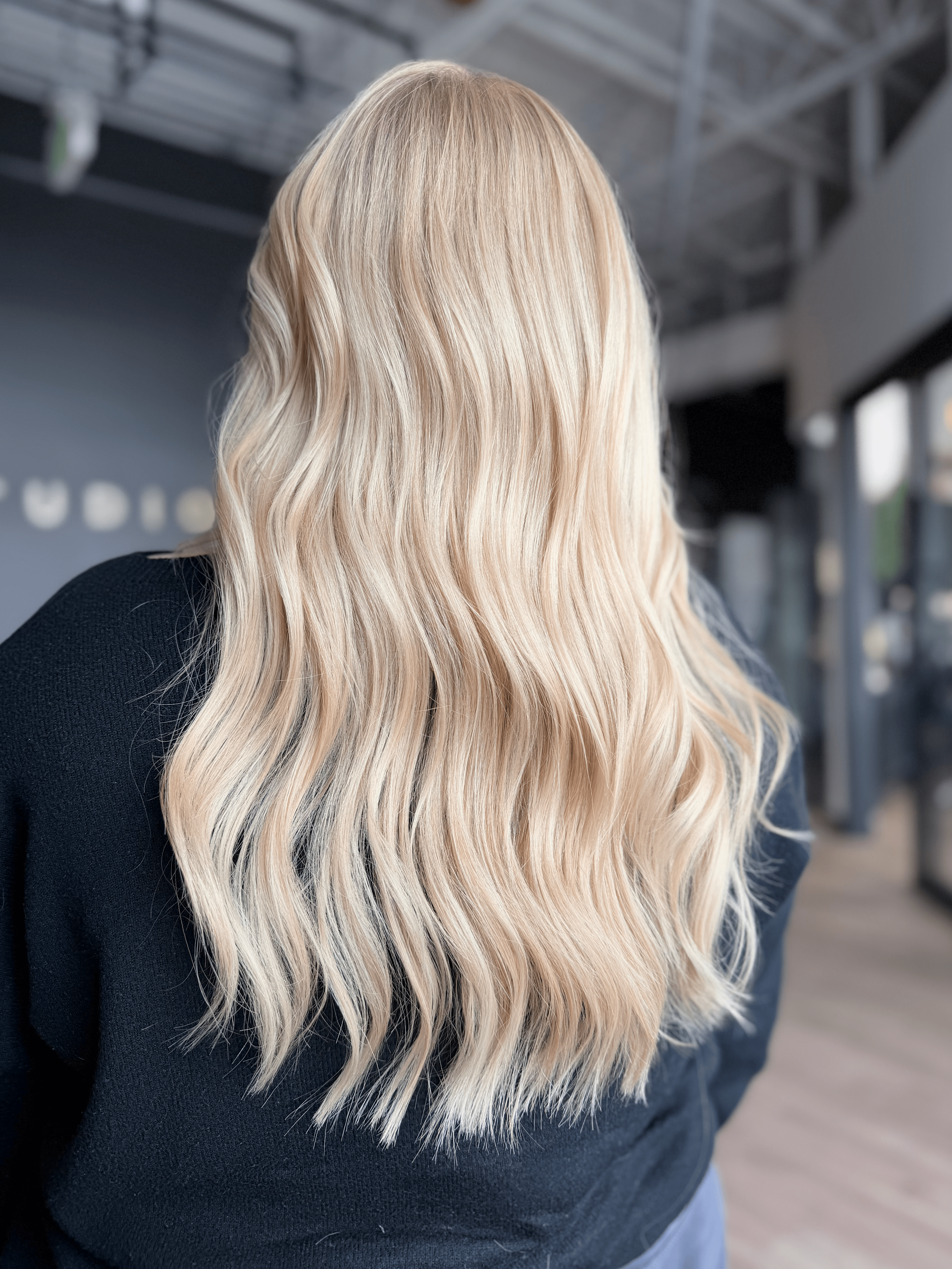 Back view of long, wavy blonde hair on a person wearing a dark top inside a modern building.