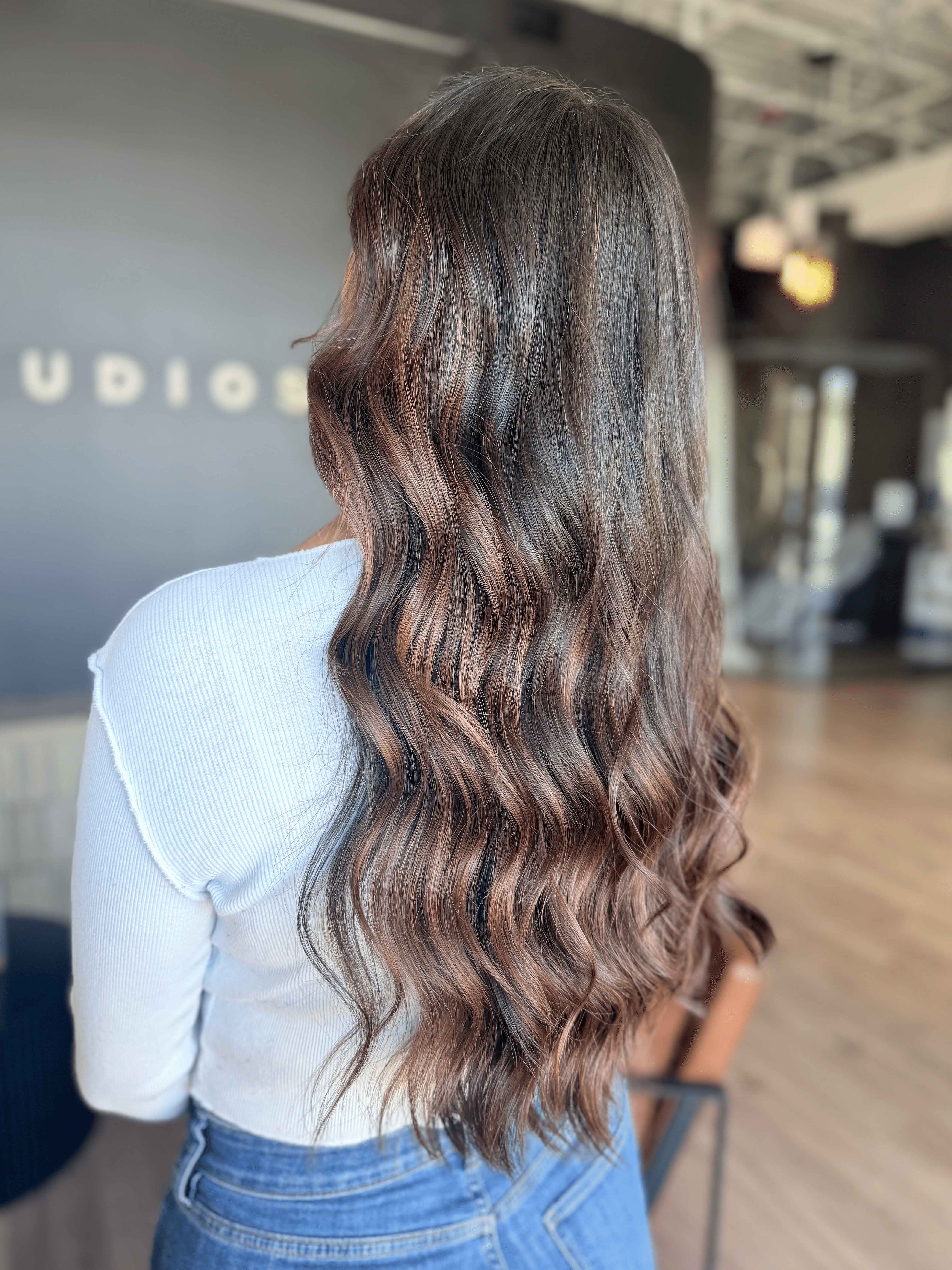 Back view of a woman with long, wavy brown hair wearing a white top and blue jeans in a modern indoor setting.