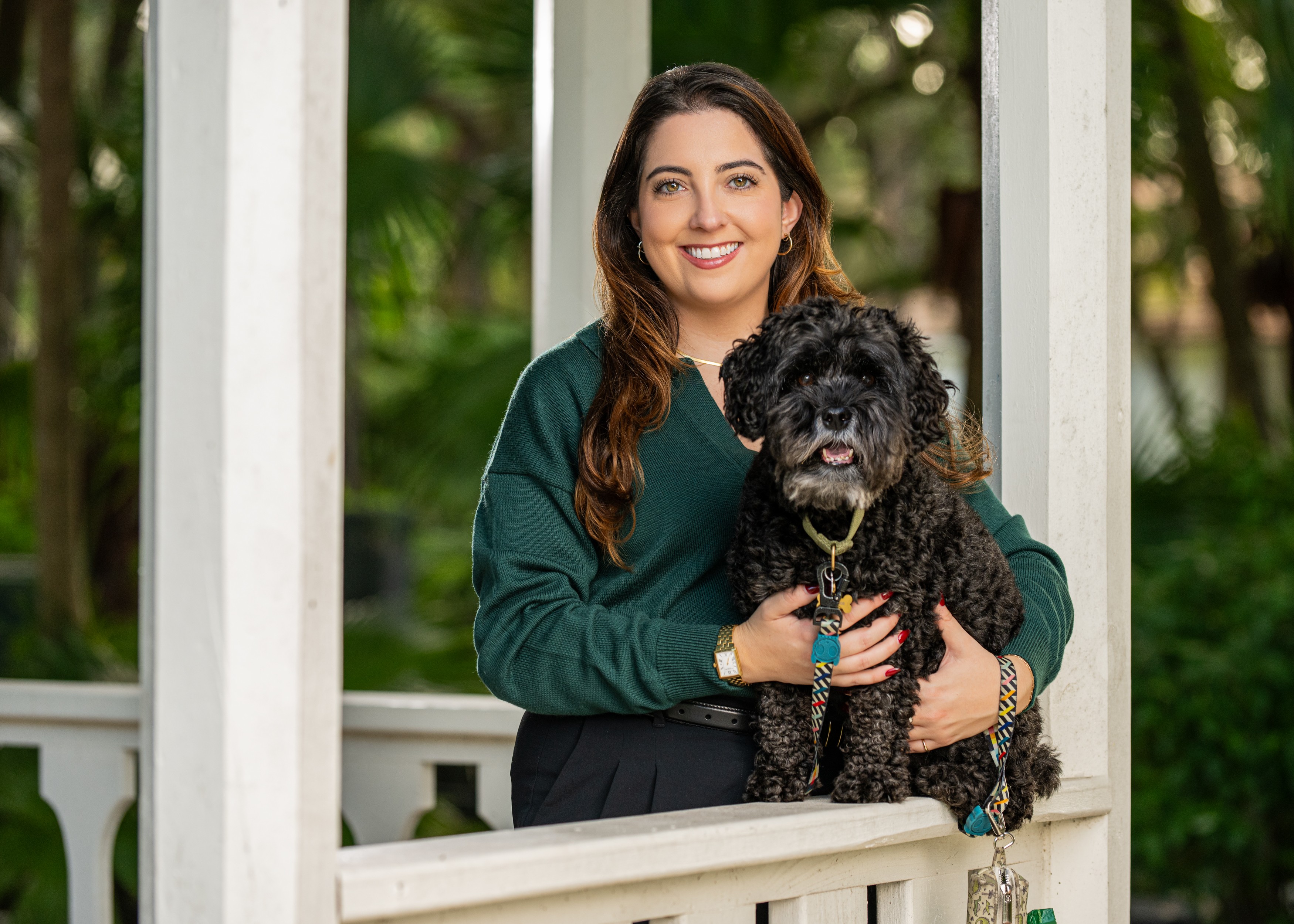 Woman smiling holding a black curly-haired dog on a porch with white railing, greenery in background.