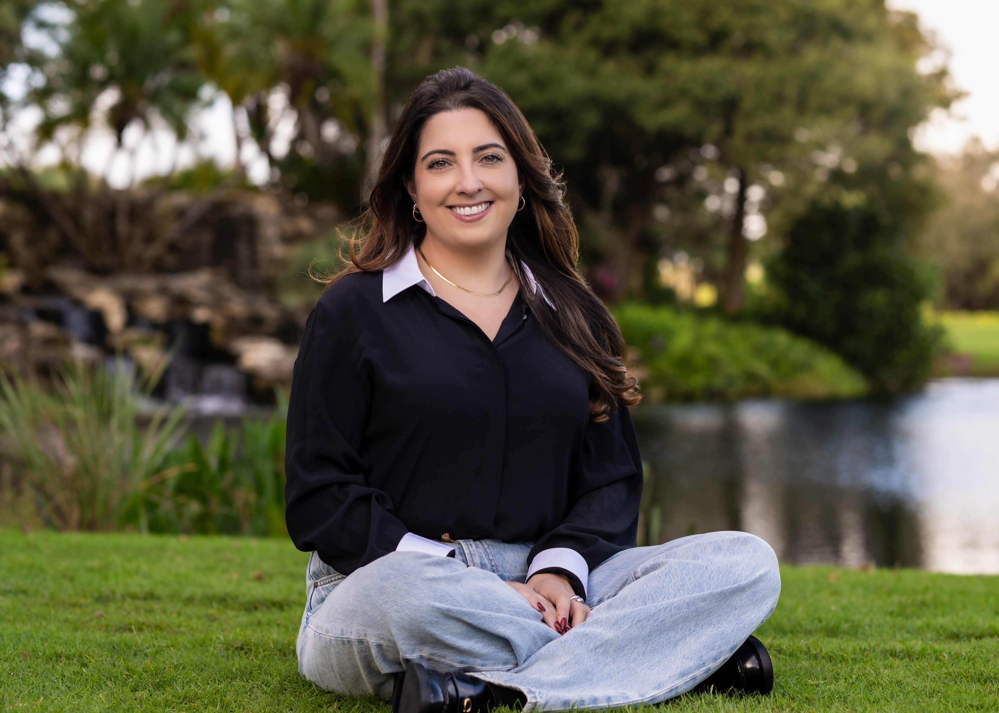 Woman sitting cross-legged on grass outdoors, smiling, with trees and water in background.