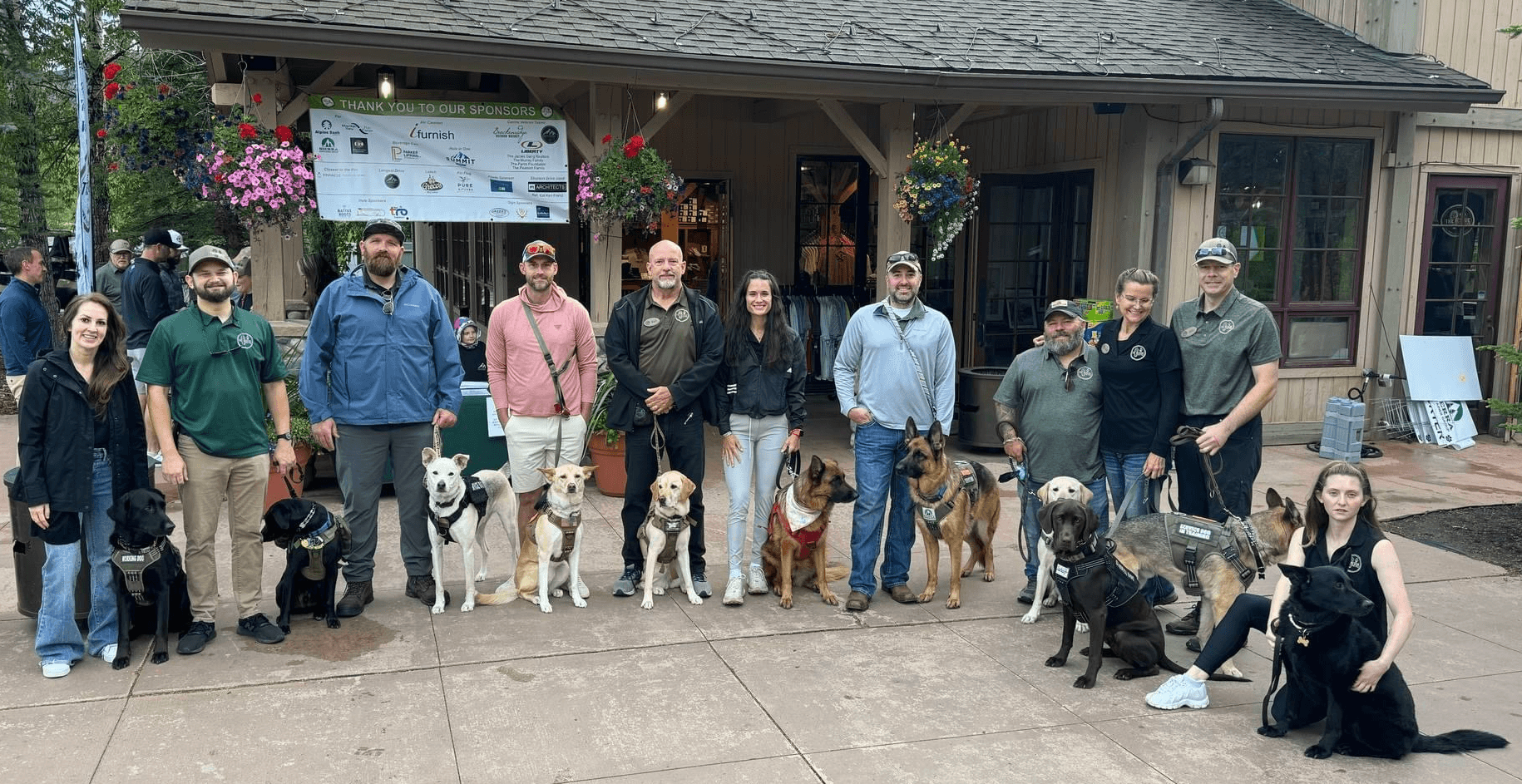 Group of people with dogs standing outside building, some sitting on ground, trees and flowers in background.