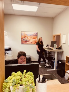 Two women in an office, one sitting at a desk and the other standing near a computer, with a painting on the wall.