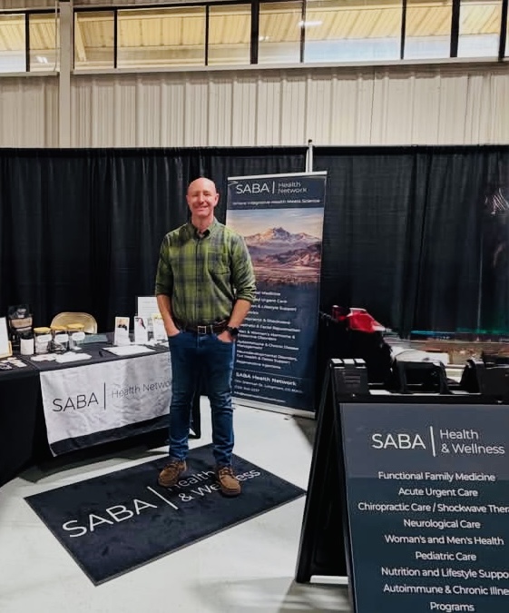 Man standing on a black carpet with SABA logo, smiling, in front of informational banners and tables at an indoor event.