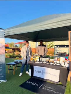 Two people standing behind a table with health products under a canopy outdoors, with a blue sky and trees in background.