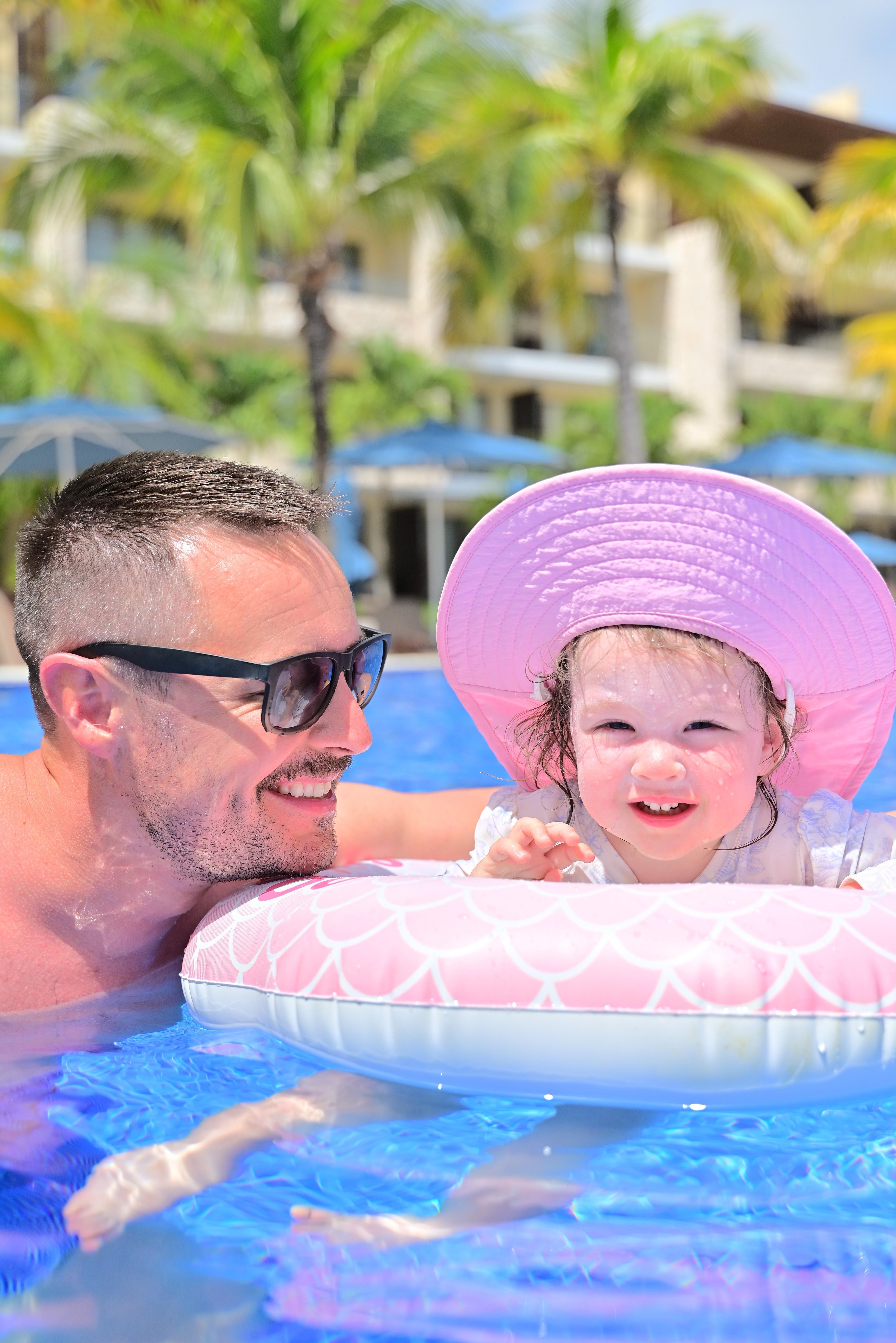 Adult man and young girl in a pool, girl wearing a large pink sun hat, smiling, with palm trees and buildings in background.