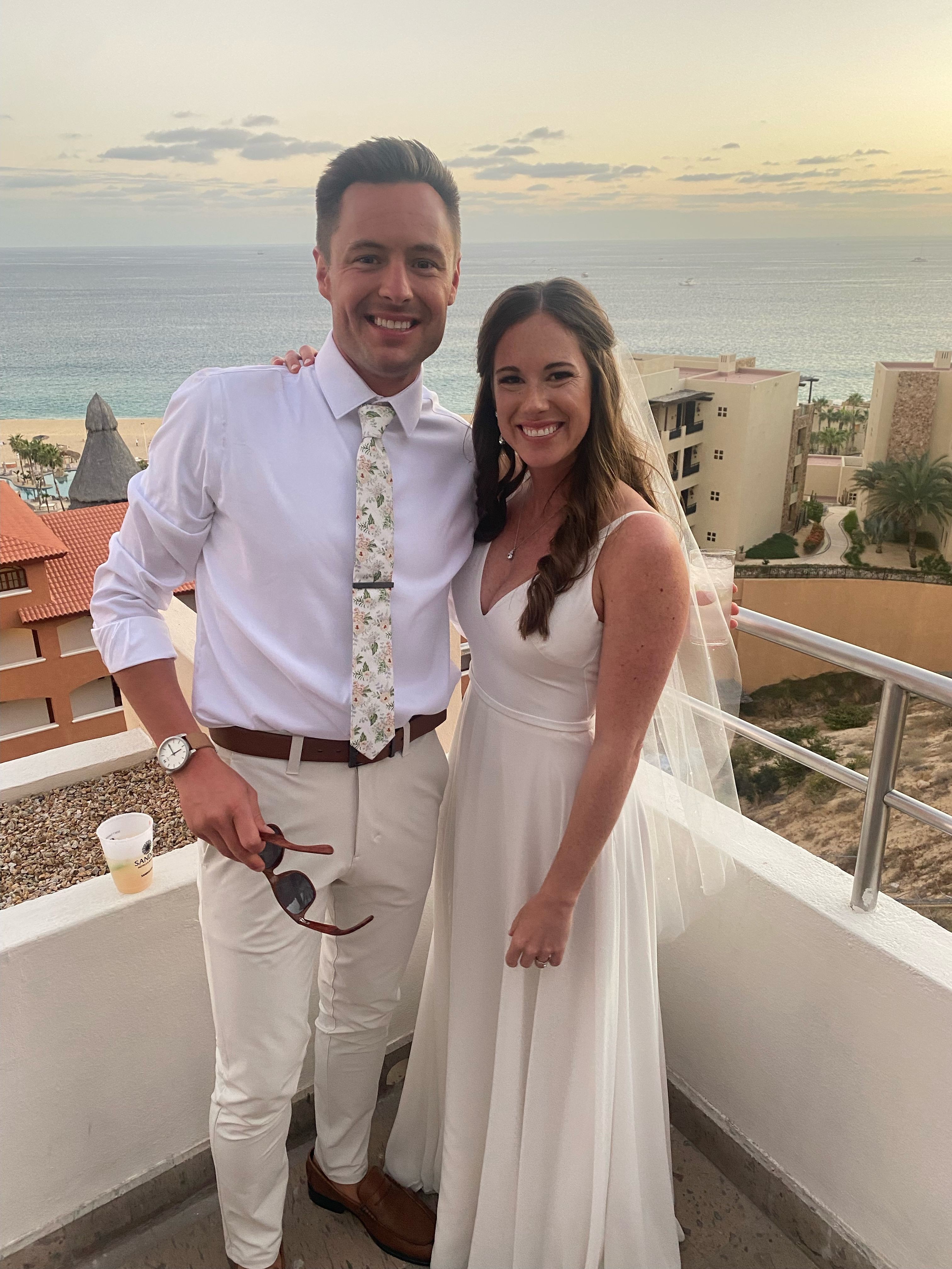 A man and woman stand on a balcony overlooking the ocean, smiling at the camera. The woman wears a white dress, and the man wears a white shirt and light-colored pants.