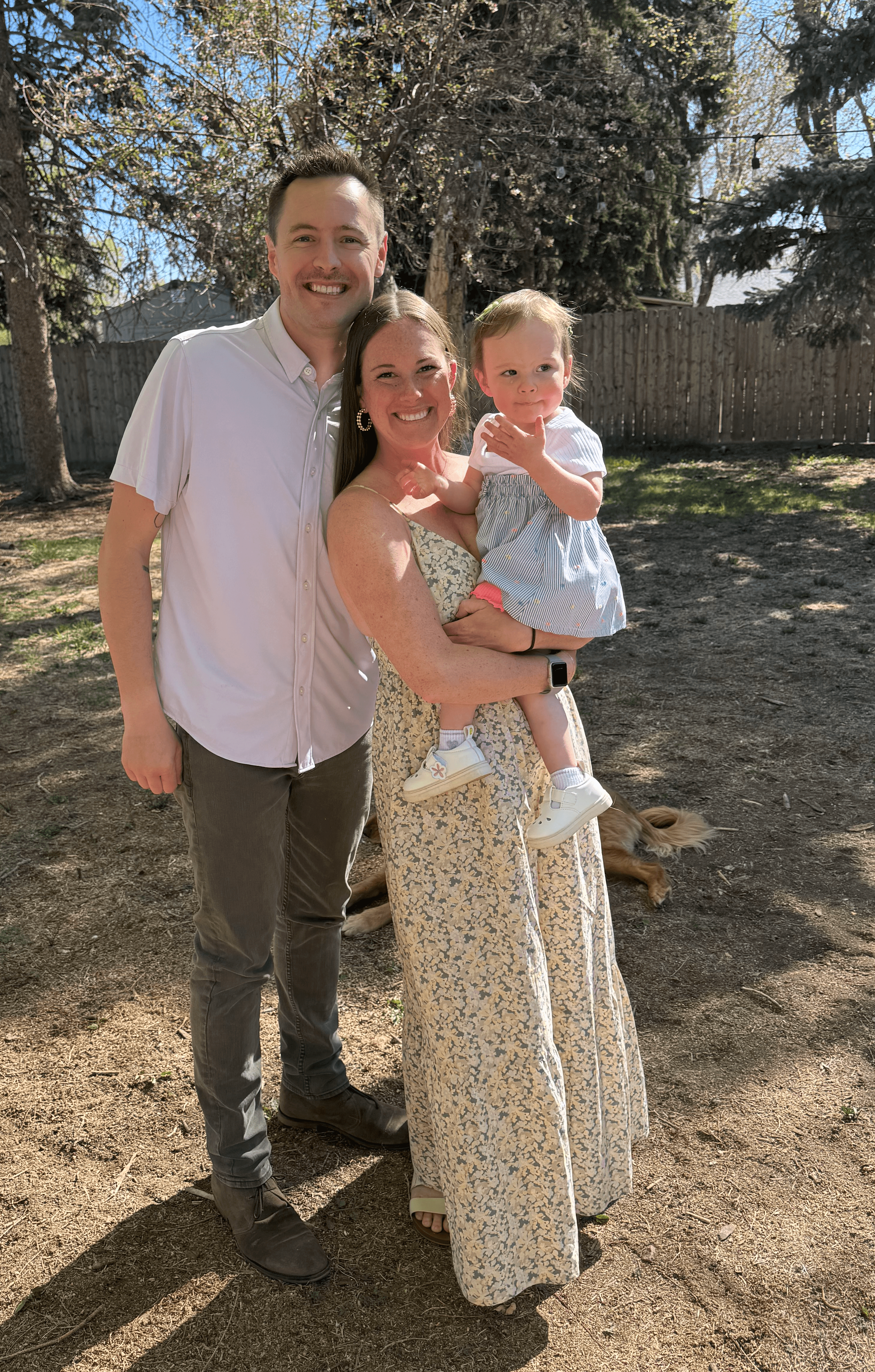 Three people outdoors: a man, a woman holding a child, all smiling, with trees and a fence in background.