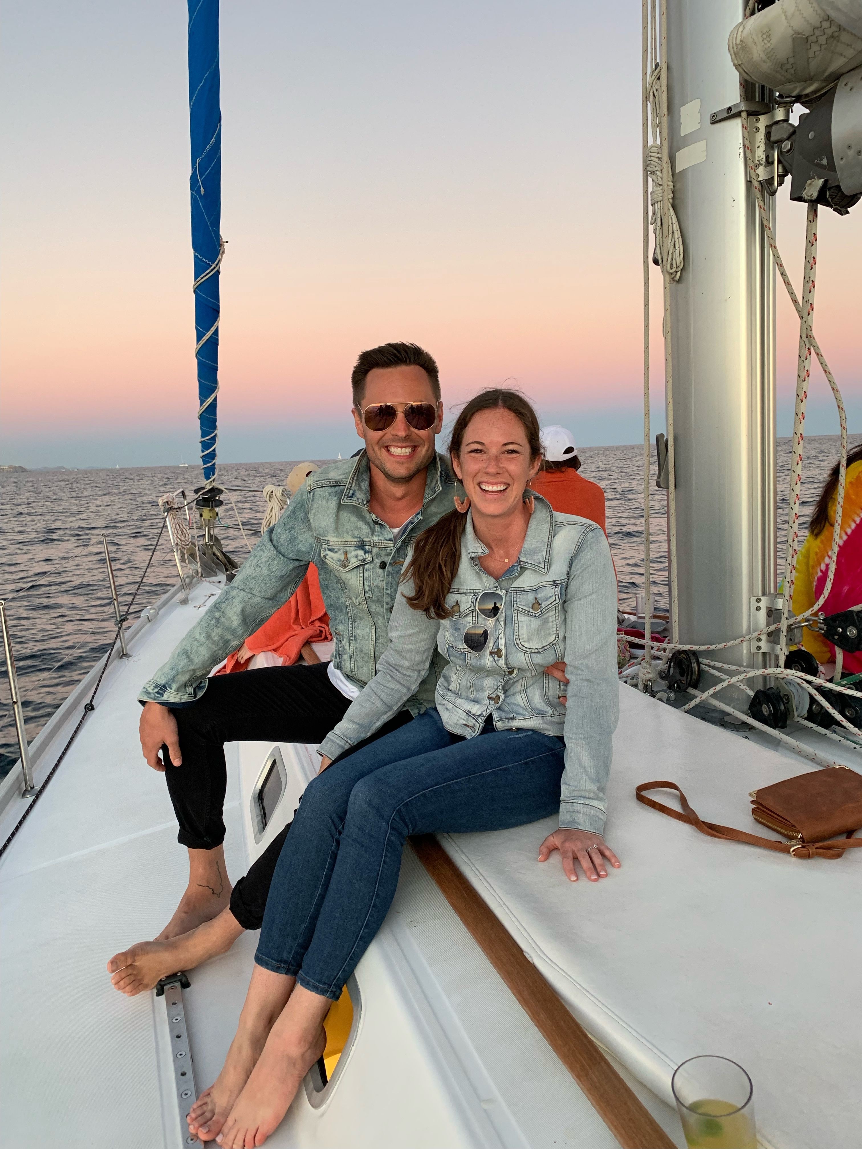 Two women sit on a boat deck with a city skyline and water in the background during sunset.