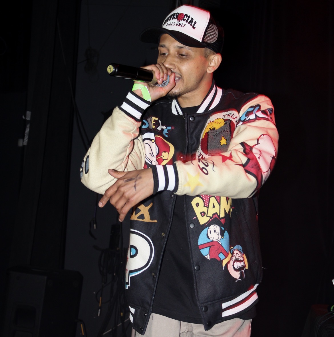 Young man singing into a microphone on stage, wearing a colorful jacket and a cap, with a dark background.