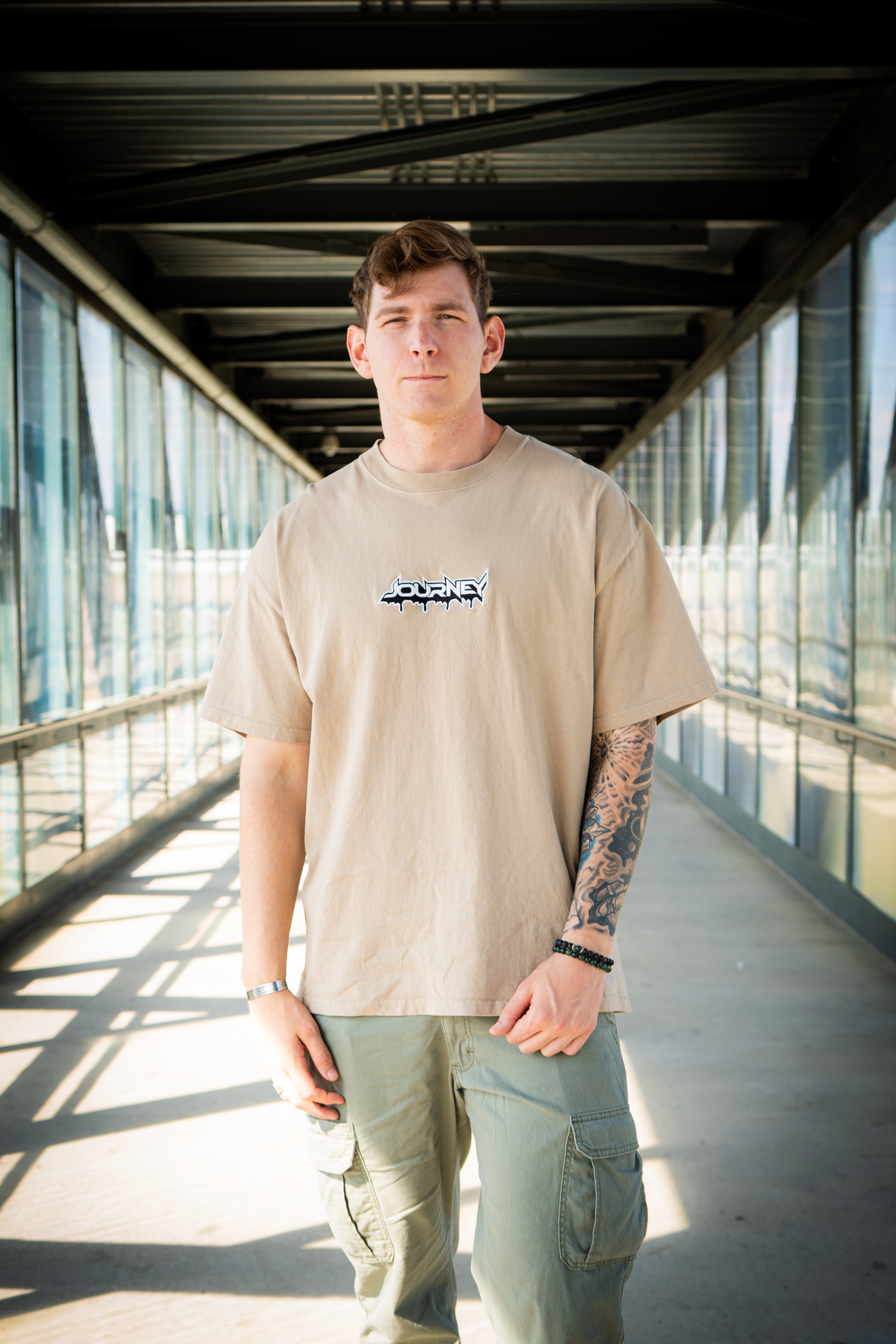Young man standing in a glass corridor with sunlight, wearing a beige t-shirt and cargo pants, with tattoos on his left arm.