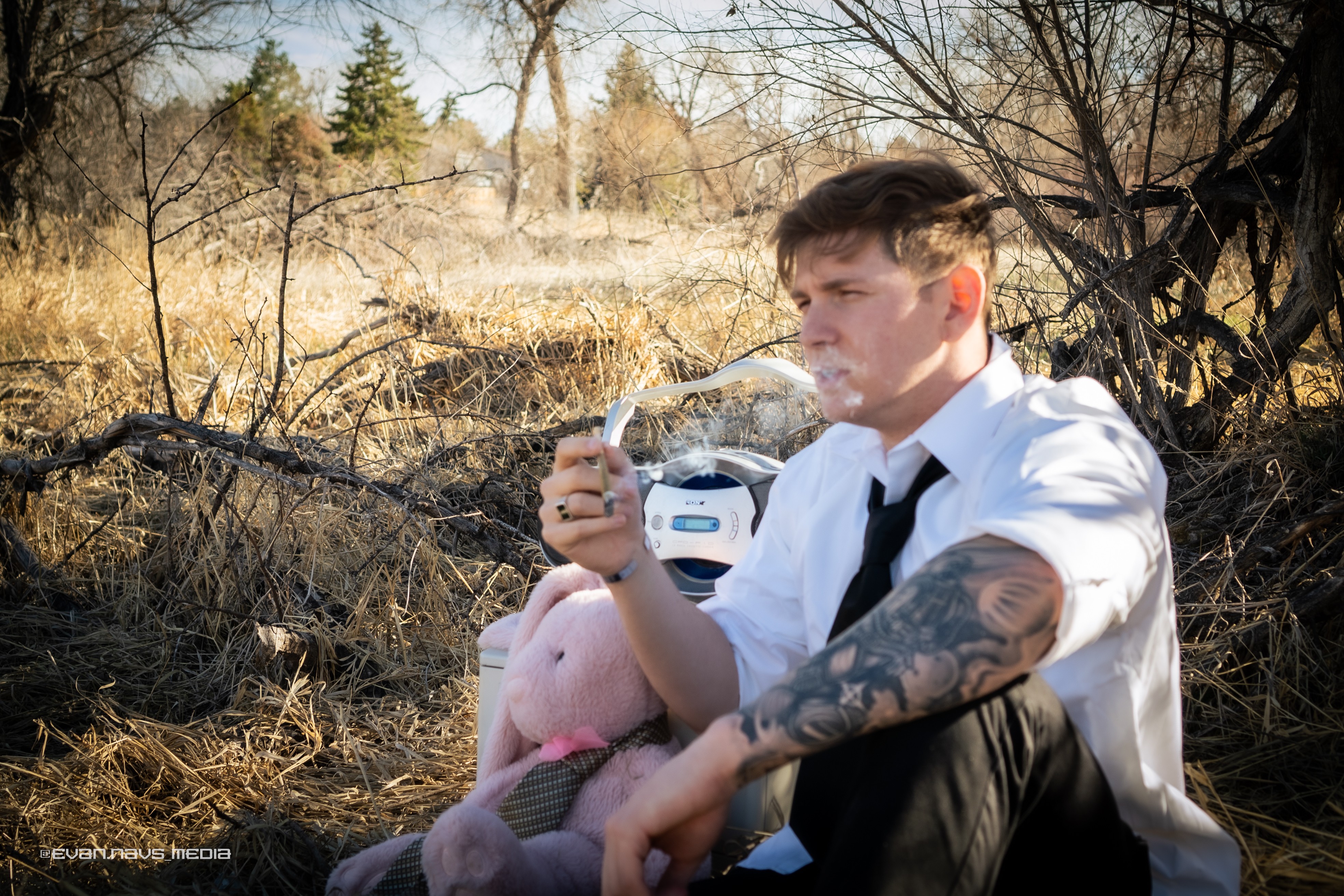 Young man with tattoos in a white shirt and black tie sitting outdoors, holding a phone, with a piglet nearby, in a natural setting.