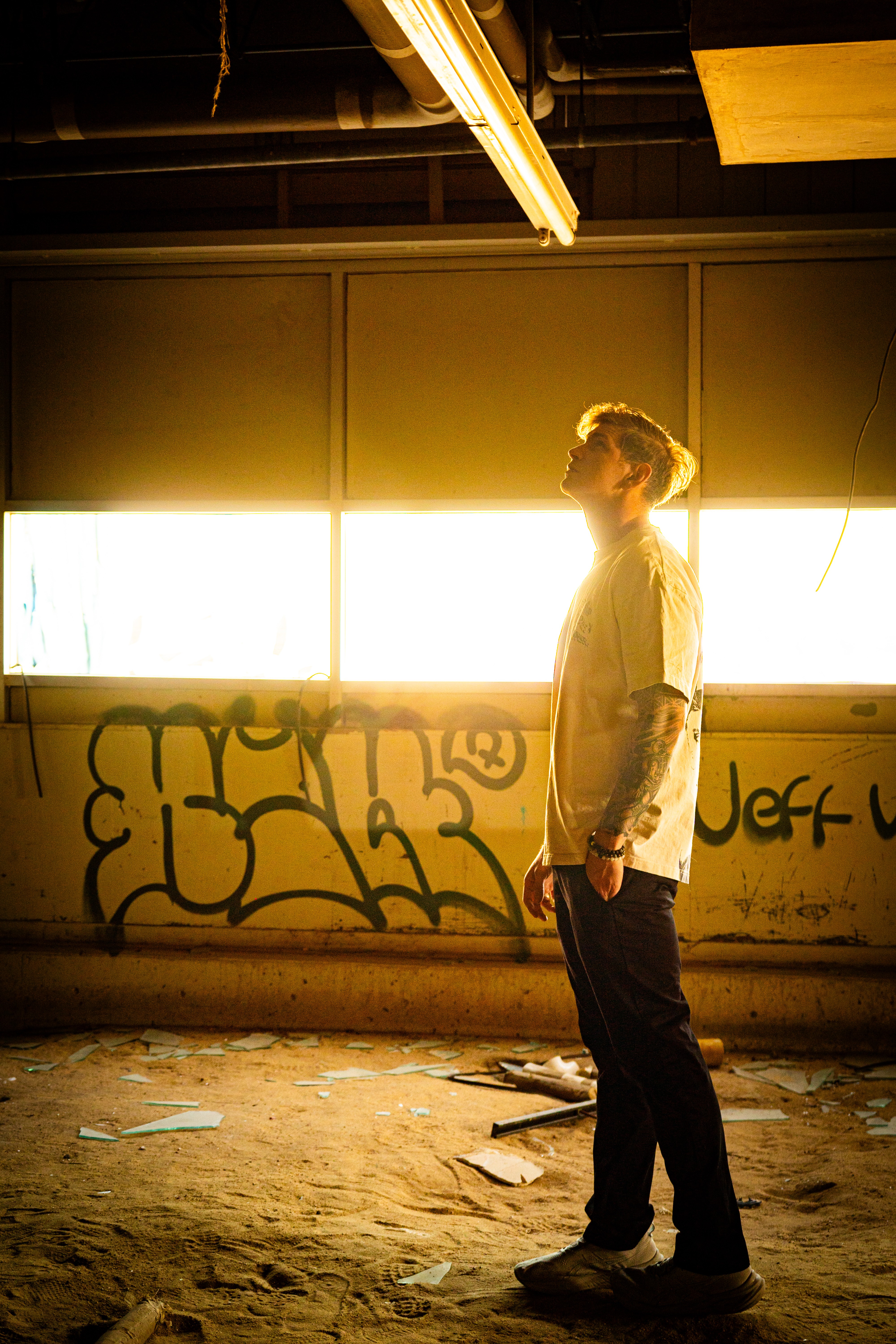 Person with hair tied back standing in an industrial space with graffiti, illuminated by bright windows behind.