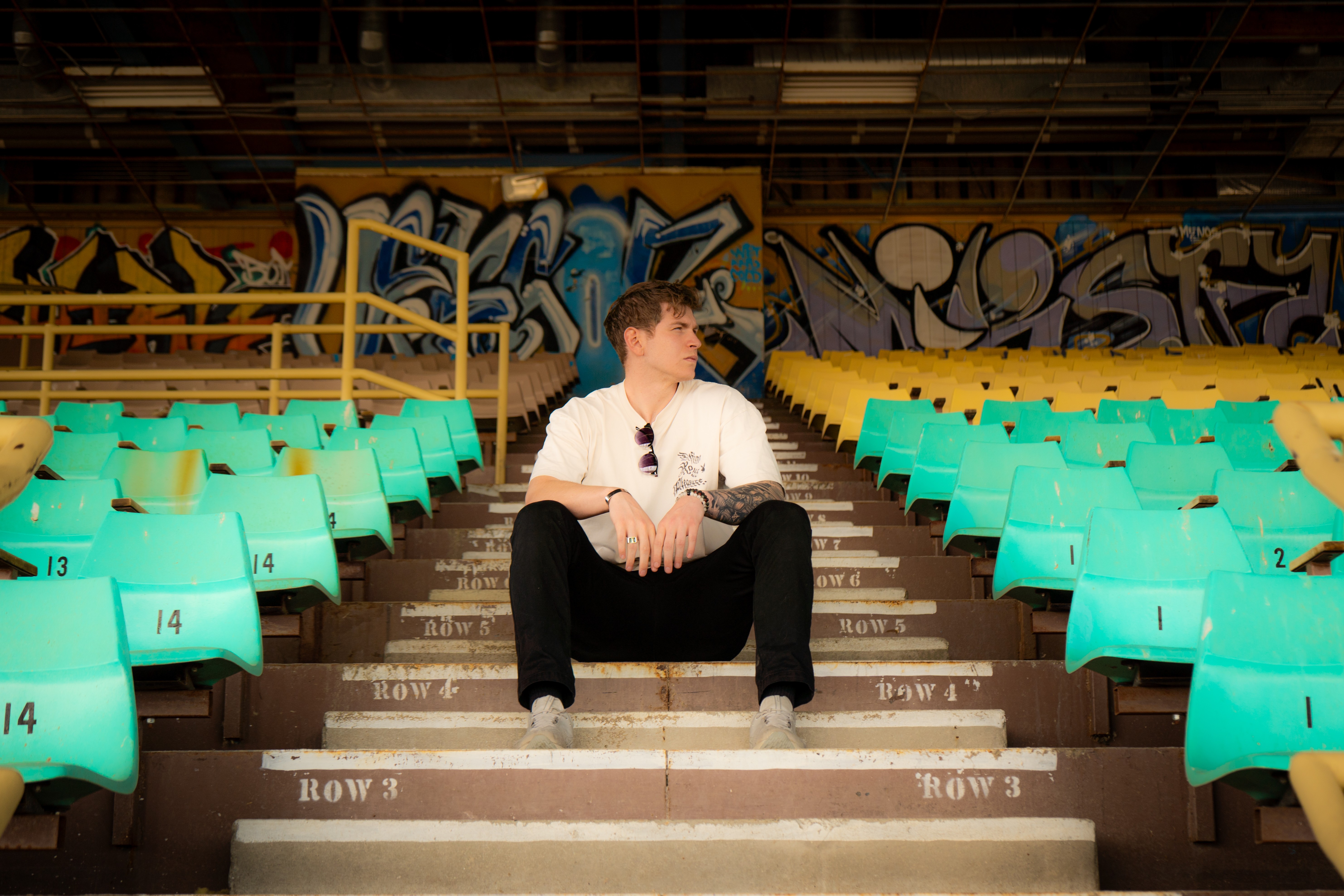 Person sitting on stadium stairs surrounded by empty teal and yellow seats, graffiti on wall behind.
