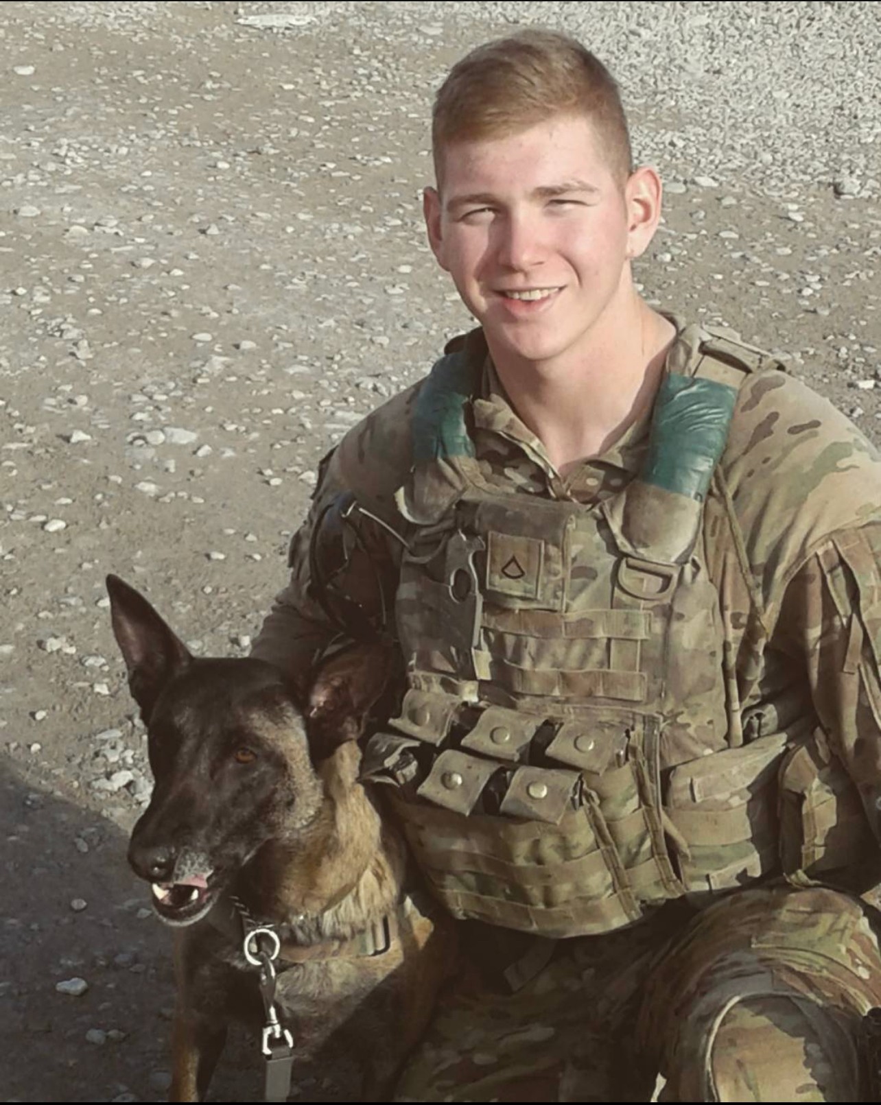 Young man in military uniform smiling with a dog on a rocky beach, background of pebbles and sand.