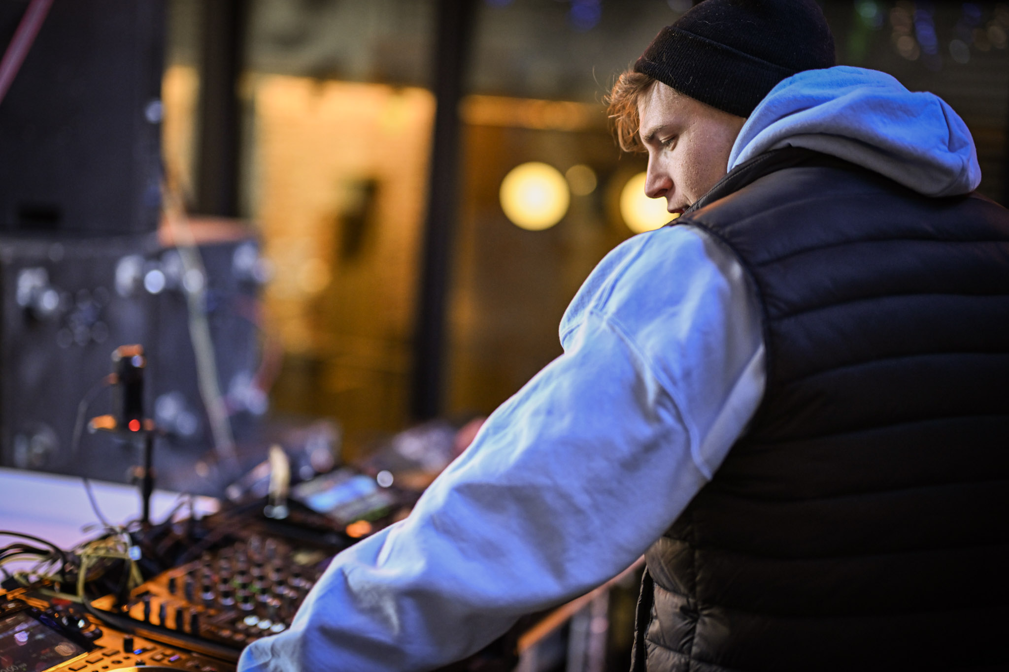 Person wearing a black beanie and blue hoodie, focused on DJ equipment in a dimly lit setting.
