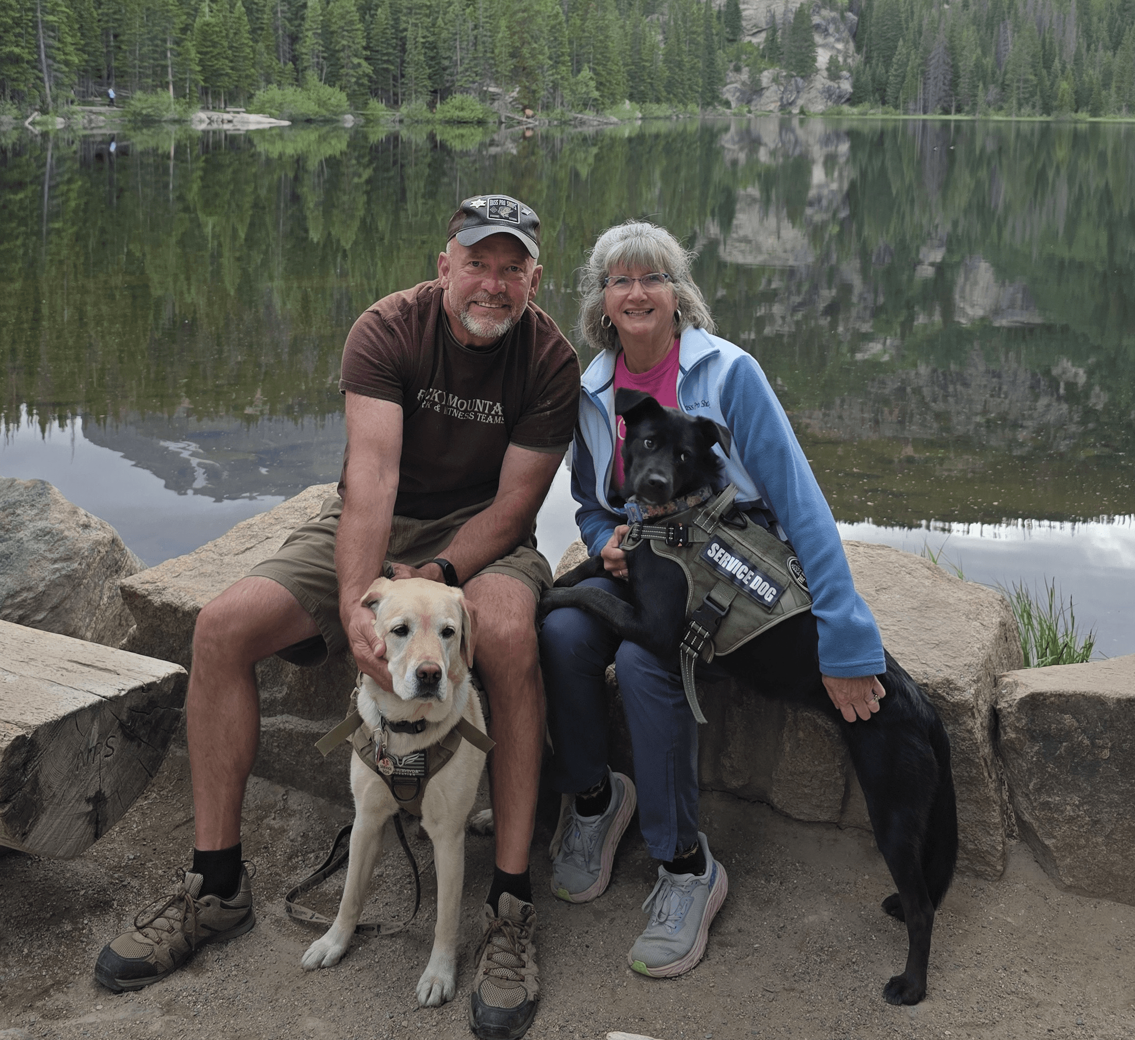 Two people and two dogs sit on rocks by a lake with trees in the background, smiling at the camera.