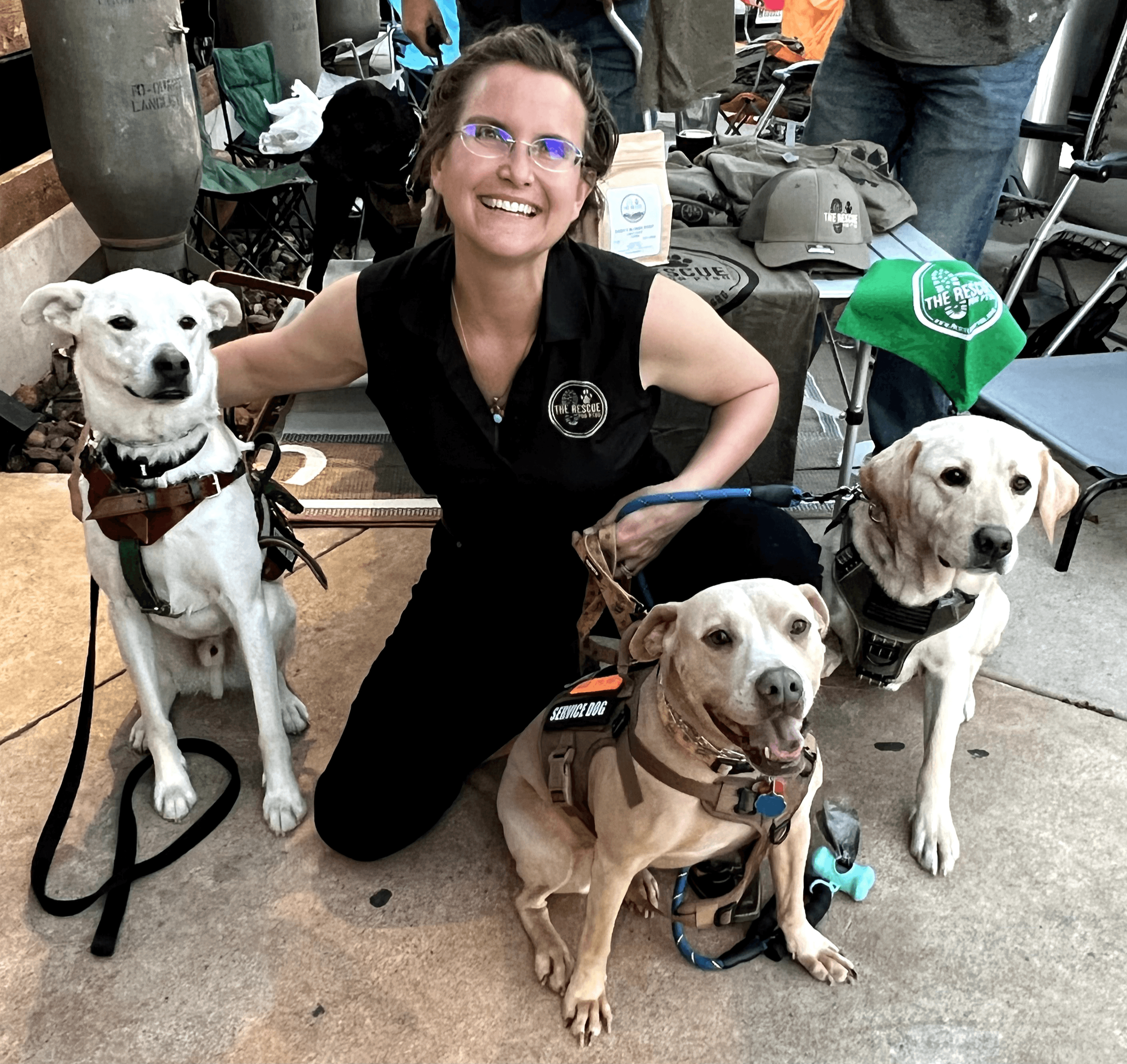 Woman kneeling with three service dogs, smiling, in an indoor setting with equipment and people in the background.