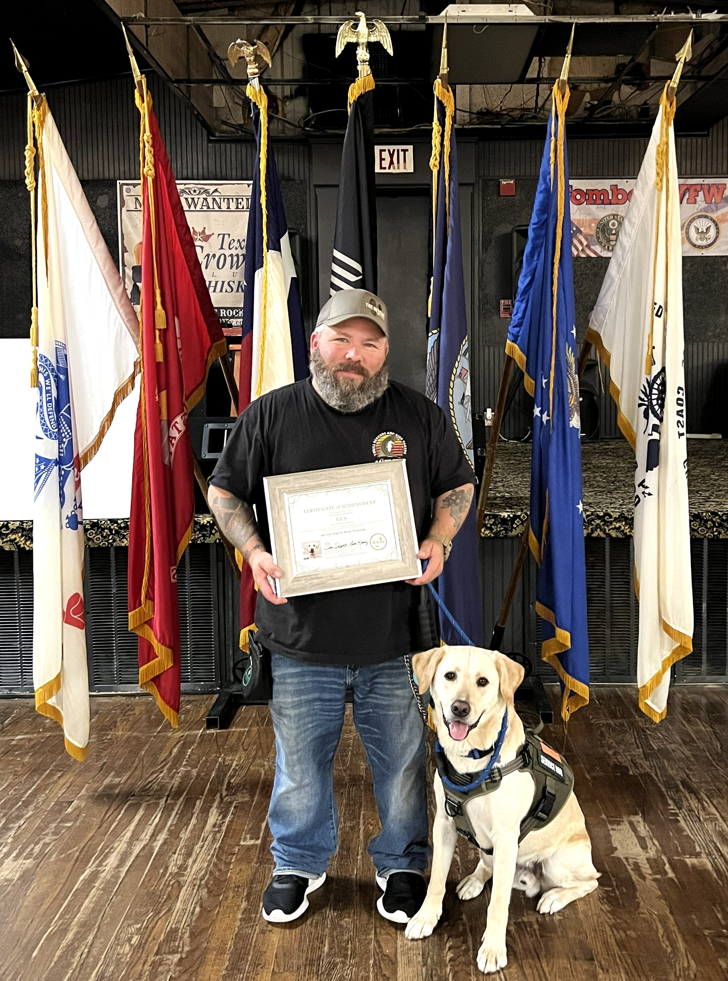 Man with a beard holding a framed certificate, sitting next to a service dog, in front of flags and banners.