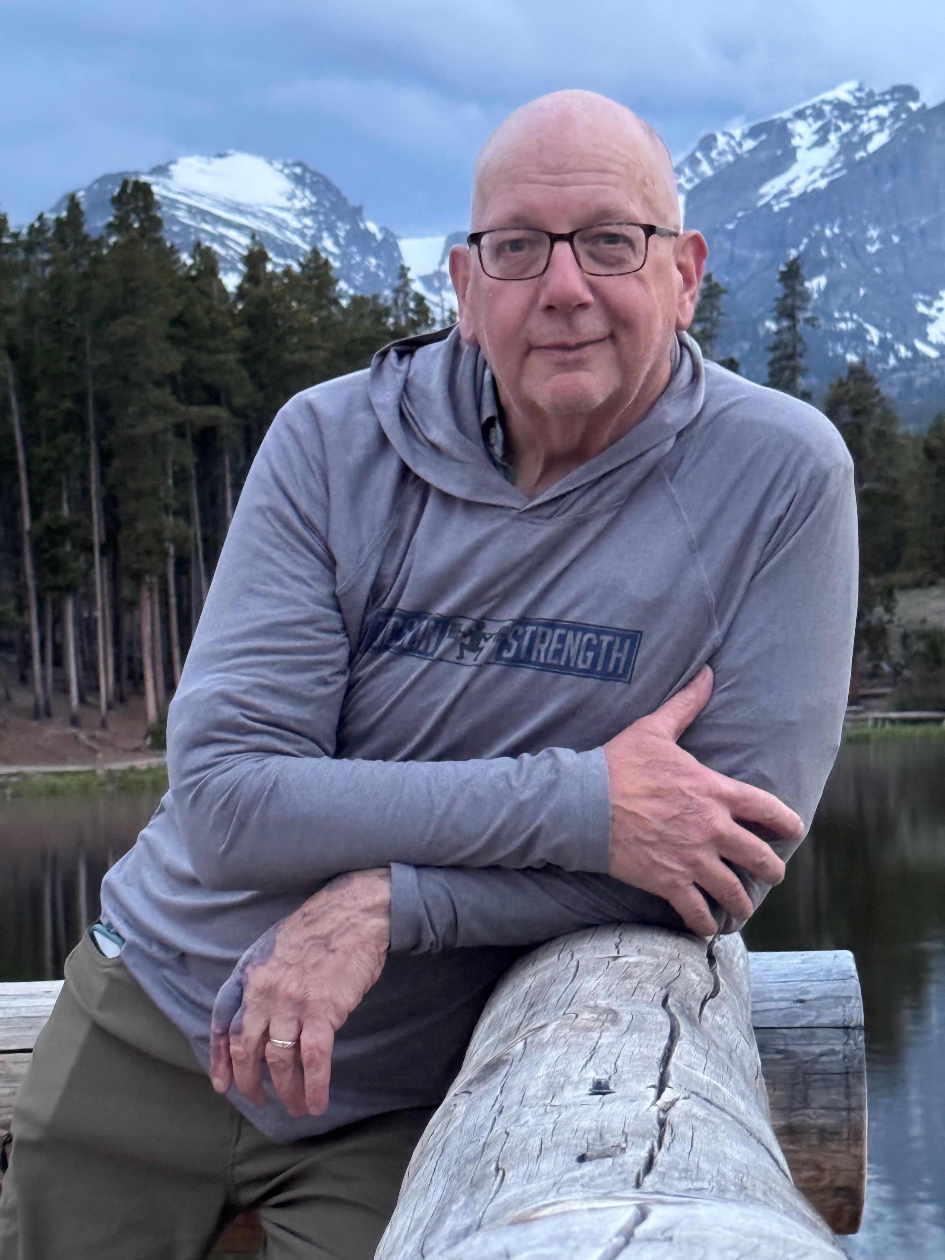 Older man with glasses and a gray hoodie sitting outdoors near trees and a log, smiling at the camera.