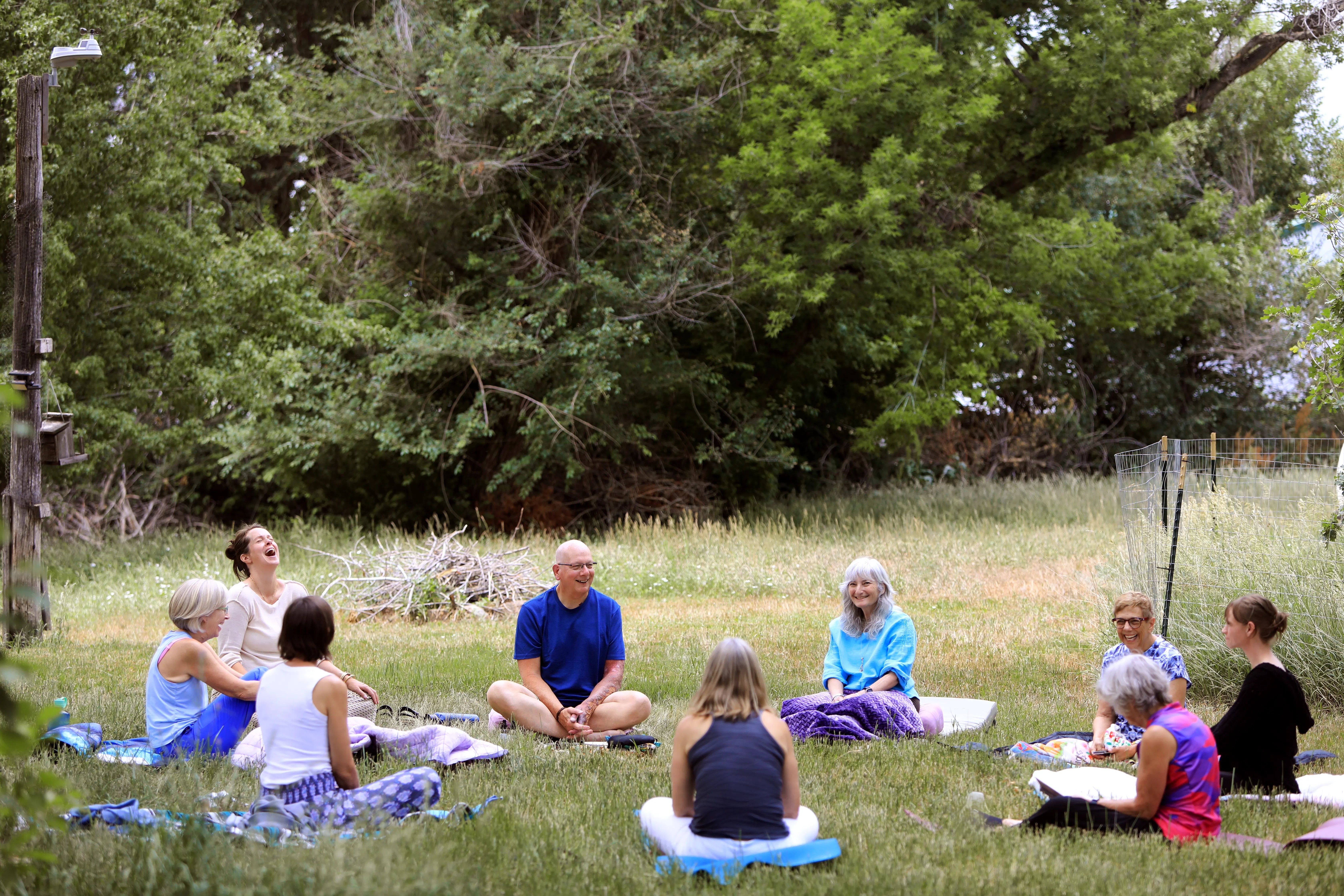 Group of people sitting cross-legged outdoors on grass, surrounded by trees, participating in a meditation or discussion session.